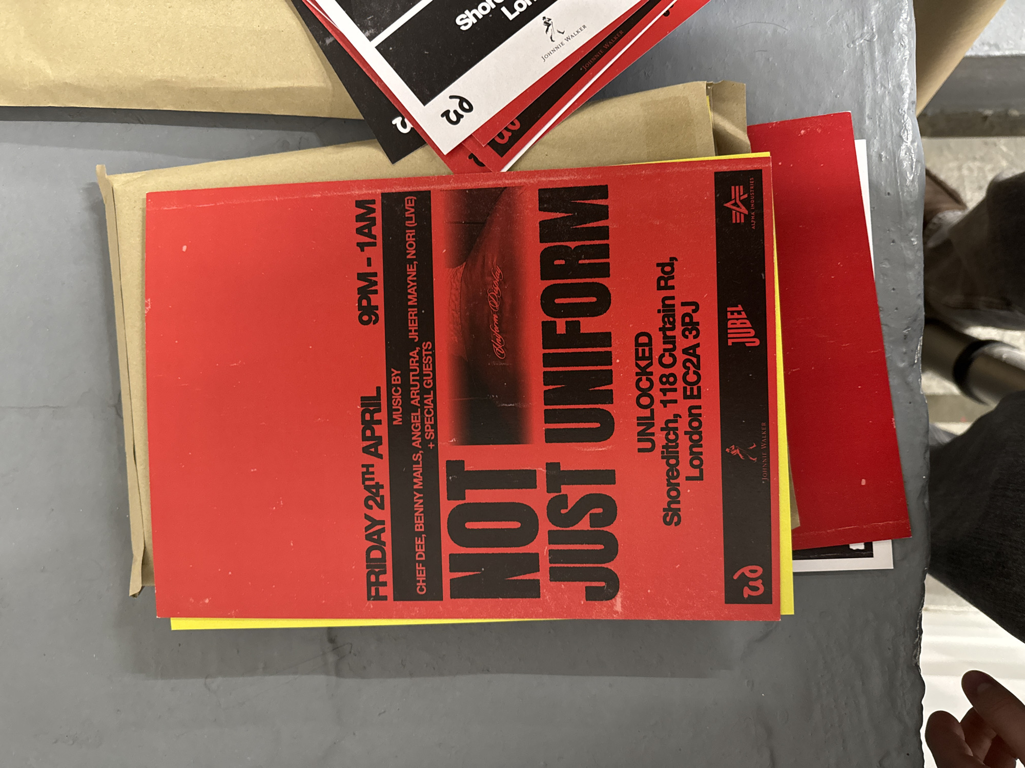 A red and black flyer for an event titled "NOT JUST UNIFORM" is centrally placed on a grey surface, likely a table or desk. The flyer, slightly worn with minor specks, advertises a music event on "FRIDAY 24TH APRIL" from "9PM - 1AM." The performing artists include "CHEF DEE, BENNY MAILS, ANGEL ARUTURA, JHERI MAYNE, NORI (LIVE)," and "SPECIAL GUESTS." The event is hosted at "UNLOCKED, Shoreditch, 118 Curtain Rd, London EC2A 3PJ."
Several logos are visible on the flyer, including "ALPHA INDUSTRIES," "JUBEL," and "JOHNNIE WALKER," along with a distinct "ud" logo. Faintly visible within the design on the flyer is the text "Uniform Display."
Beneath the main red flyer, parts of other flyers and cards are visible, predominantly in black, white, and yellow. These also feature the "JOHNNIE WALKER" logo and partial text such as "Sho" and "Lo," likely referring to "Shoreditch" and "London." A brown, envelope-like object is partially visible on the left side, under the flyers. A hand is partially visible in the bottom right corner of the frame. The overall scene suggests an indoor setting, possibly an office or event space, with materials related to an upcoming event.