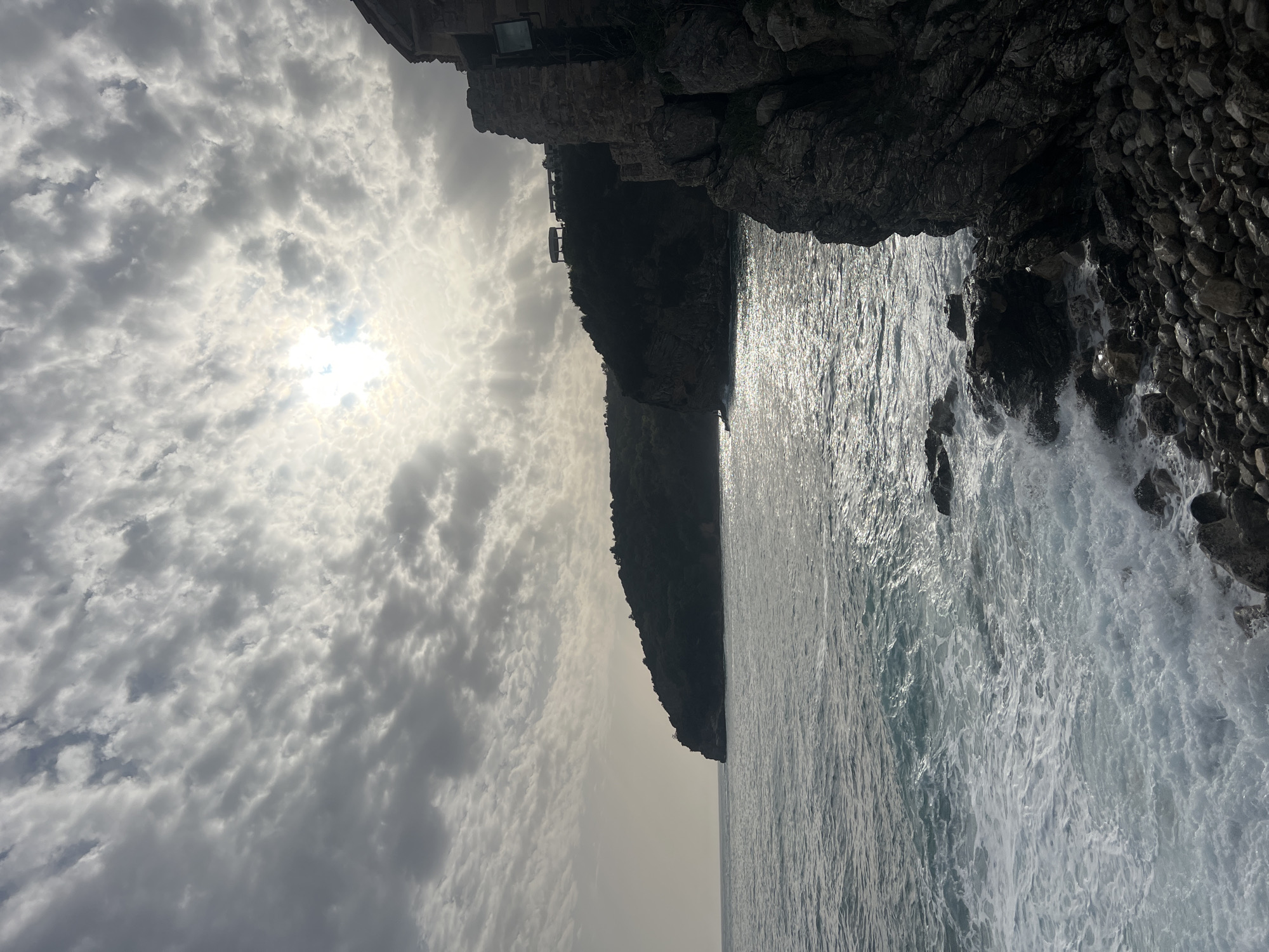 The image captures a coastal scene in Budva, Montenegro. A rocky cliff extends into the sea, with waves crashing against the base. A building is perched on the edge of the cliff. Above, a cloudy sky obscures the sun.