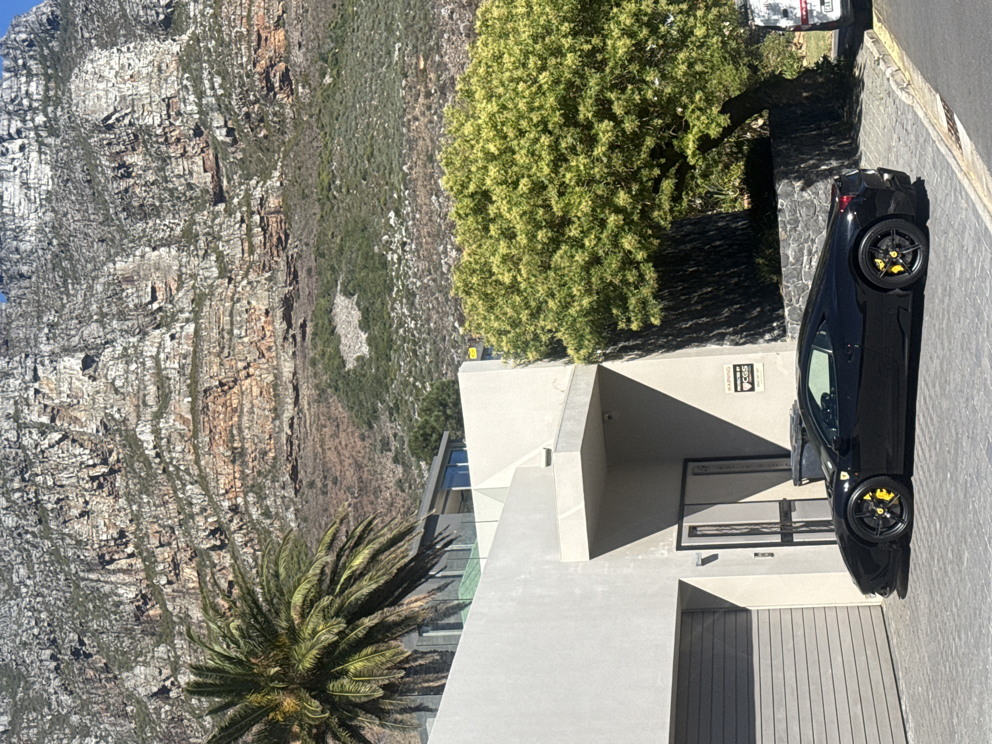 The image shows a modern house at the foot of Table Mountain in Cape Town, South Africa. A black sports car is parked in the driveway, with visible yellow brake calipers. A palm tree grows near the house, and a deciduous tree is planted between the property and the neighboring street. The building is a concrete structure with modern architectural design. The ground is paved with paving stones.