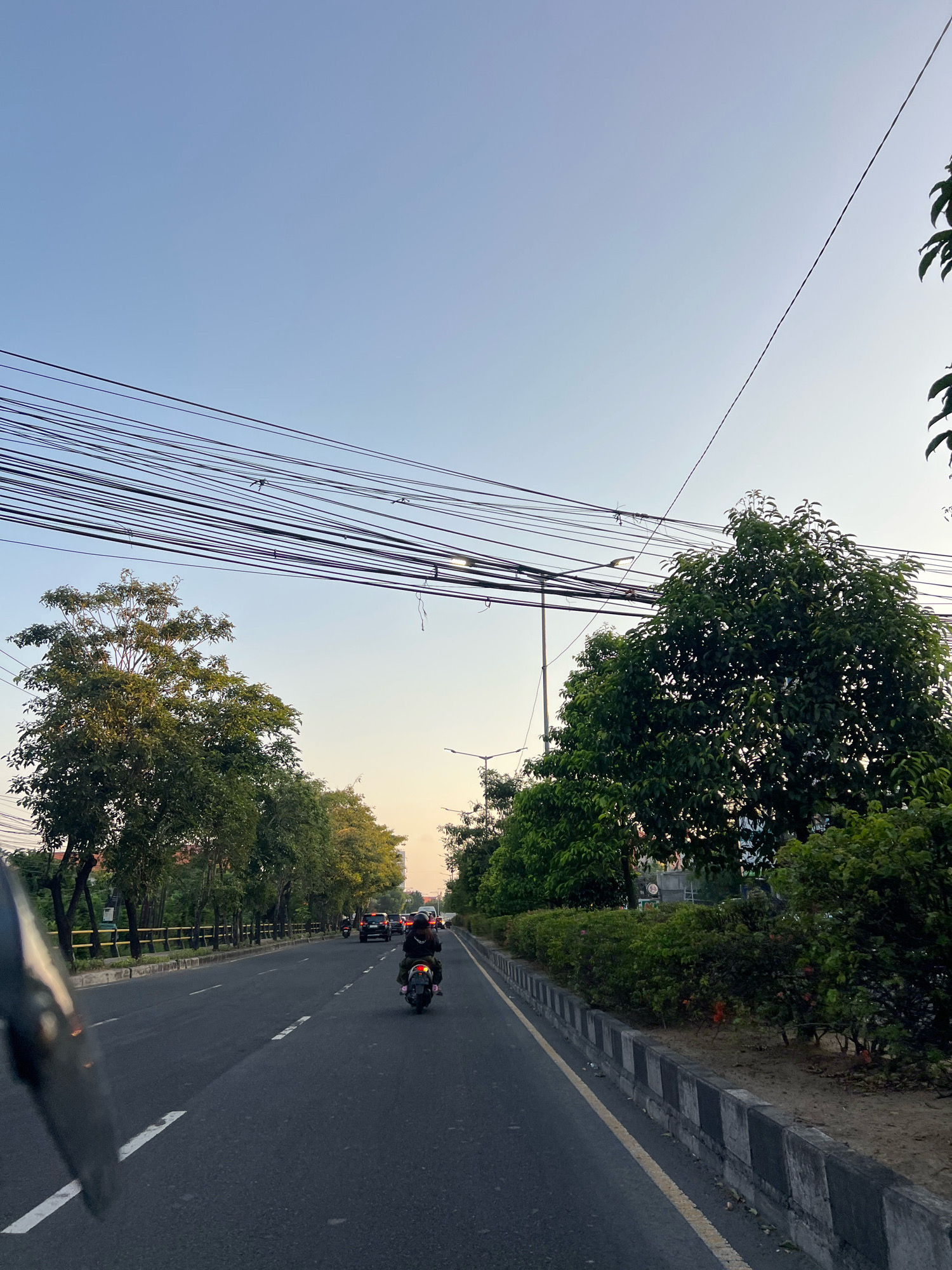 The image depicts a street scene in Kuta, Indonesia. The perspective is from the front of a motorcycle, looking forward. The road is asphalt with white and yellow lane markings. On the right side of the road, there is a concrete barrier with black and white checkered patterns, followed by a strip of sand and dense green bushes and trees. On the left side of the road, there is a sidewalk with a yellow metal fence and a row of tall trees with green foliage. In the distance, several cars and another motorcycle are visible on the road. A person is riding the motorcycle in the foreground, facing away from the camera. The sky above is a clear, pale blue, with a large bundle of black power lines stretching across the upper portion of the frame. The lighting suggests it is either early morning or late afternoon, with a soft, diffused light.
