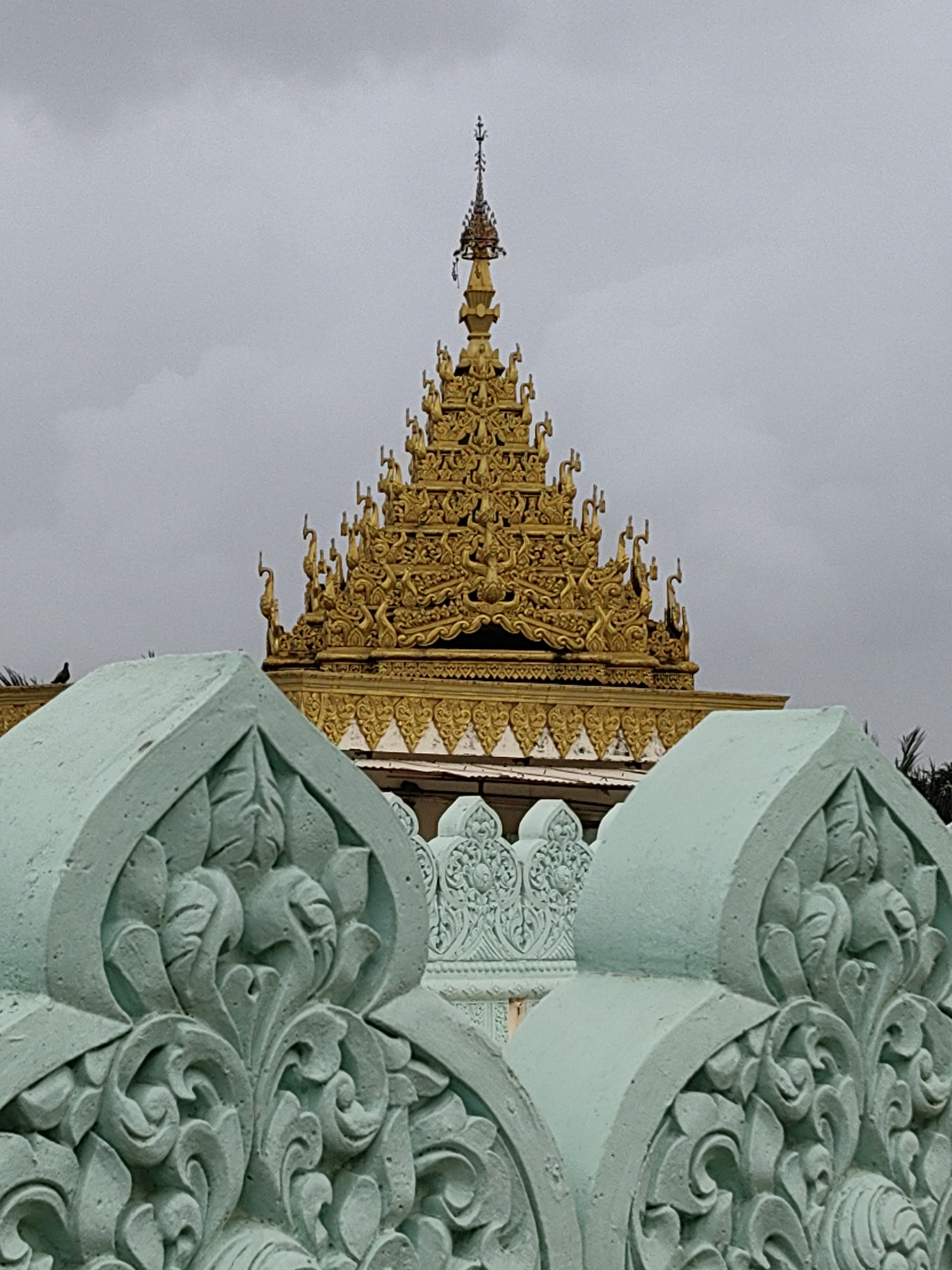 The image captures a close-up view of ornate decorative structures in the foreground, appearing to be part of a fence or balustrade. These structures are rendered in a pale, mint-green color and feature intricate, carved floral and scrollwork patterns. Behind this detailed foreground, a golden tiered roof with elaborate ornamentation is visible, likely belonging to a temple or religious building. The roof is adorned with small figures and decorative elements, characteristic of Southeast Asian architectural styles. The sky above is overcast and grayish, suggesting a cloudy day. The overall scene evokes a sense of detailed craftsmanship and cultural heritage. While the image does not provide explicit landmarks to pinpoint the exact location in Mumbai, the architectural style of the golden structure is reminiscent of Buddhist or Hindu temples found in India, and specifically, this style is common in and around Mumbai.