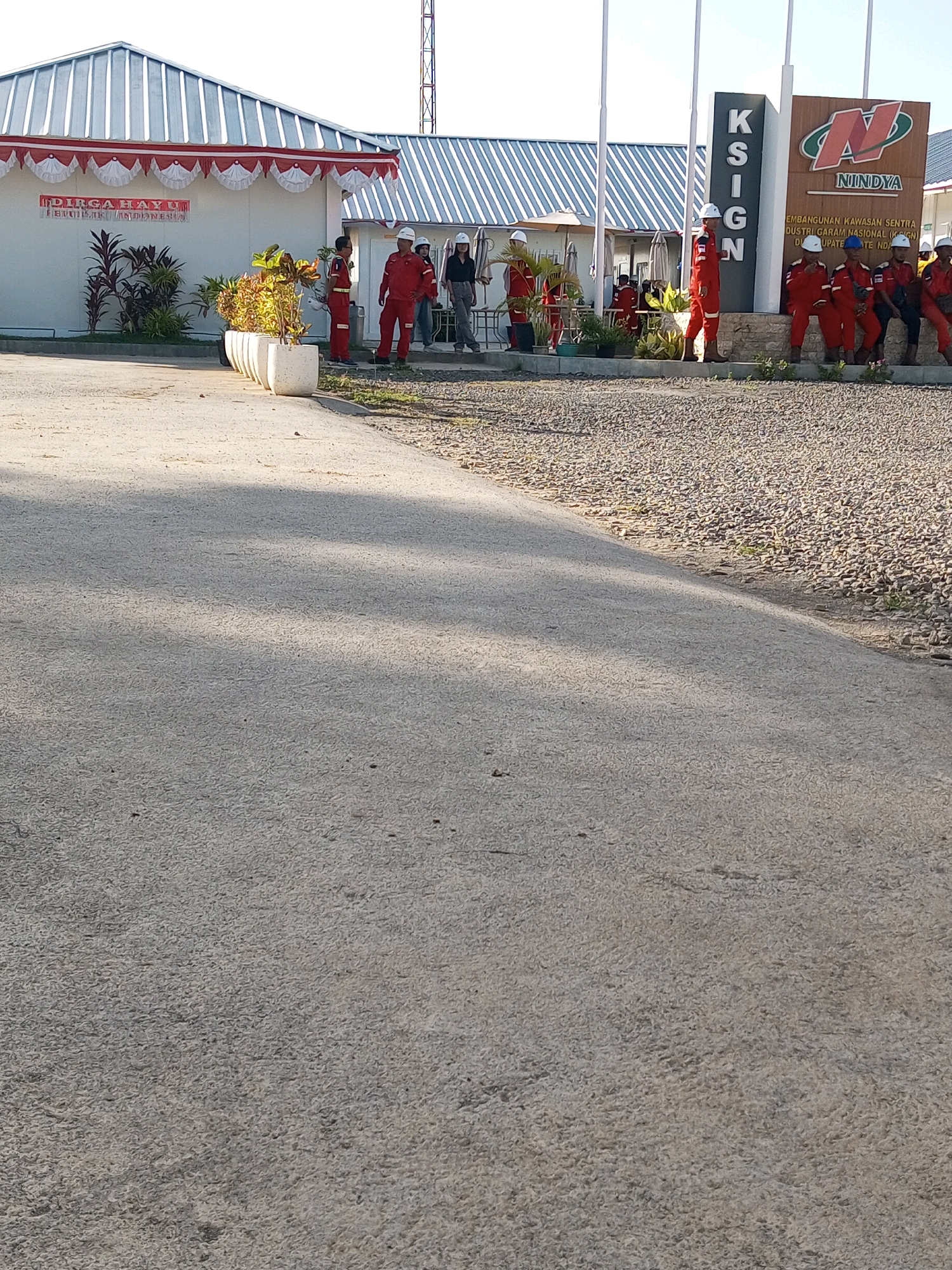 The image captures an outdoor scene at the Nindya Karya construction site in Matasio, Indonesia. A group of approximately ten individuals, most wearing red work overalls and white hard hats, are present. Some are standing, while others are seated on a low wall or the ground near a large sign. The sign, prominently featuring the "Nindya" logo, also displays text indicating "PEMBANGUNAN KAWASAN SENTRA INDUSTRI GARAM NASIONAL (KSIGN) DUPATE ND".
To the left, a white building with a corrugated metal roof is visible. Red and white festive banners are draped along the eaves of this building, suggesting a possible celebration or event. Several potted plants, including some with red and green foliage, are arranged in front of the building. The ground in the foreground is paved with asphalt, transitioning to a gravel-covered area on the right side of the image. A tall, slender structure, likely a radio tower or antenna, can be seen in the background against a clear sky. The overall impression is of a work site during daylight hours, with personnel present.
