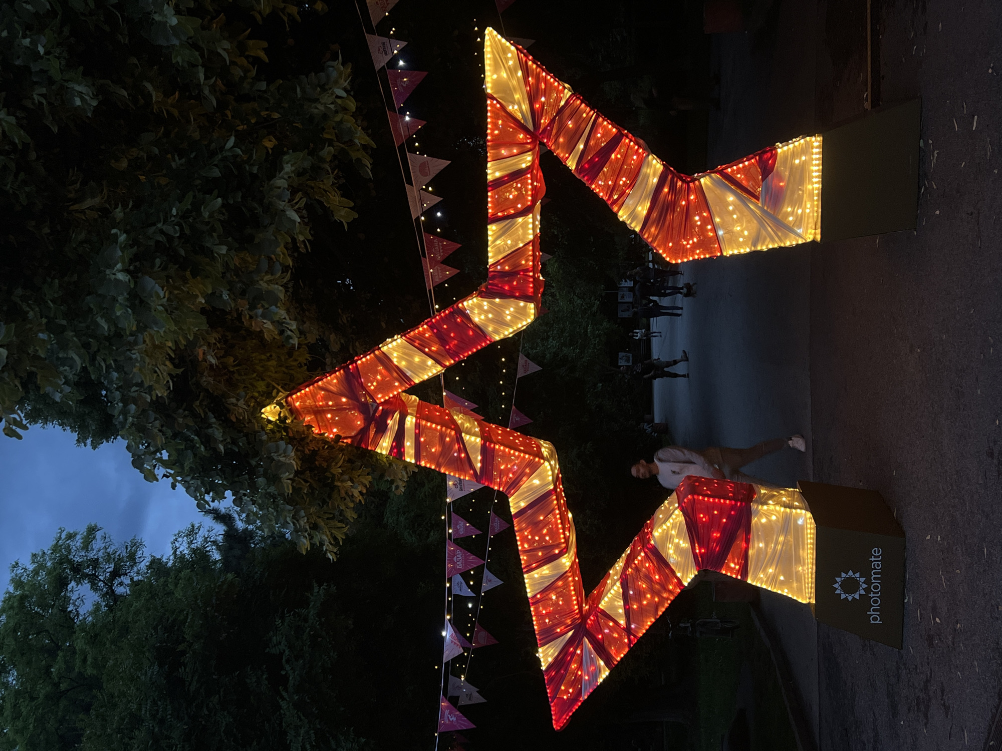 The image shows a large, illuminated zigzag structure in a park setting, likely at night. The structure is decorated with alternating sections of red and white fabric, adorned with lights. It is supported by two stands, one of which has the word "photomate" printed on it. A string of triangular flags hangs above the zigzag. In the background, there are indistinct figures of people and trees. The sky is visible in the upper left corner, showing the transition from dusk to night.
Based on the style of the decoration and the presence of Cyrillic characters in the surrounding area (evident in similar images found online), the location is likely in Plovdiv, Bulgaria. The image potentially captures an outdoor installation or art piece set up in a park for an event or festival.