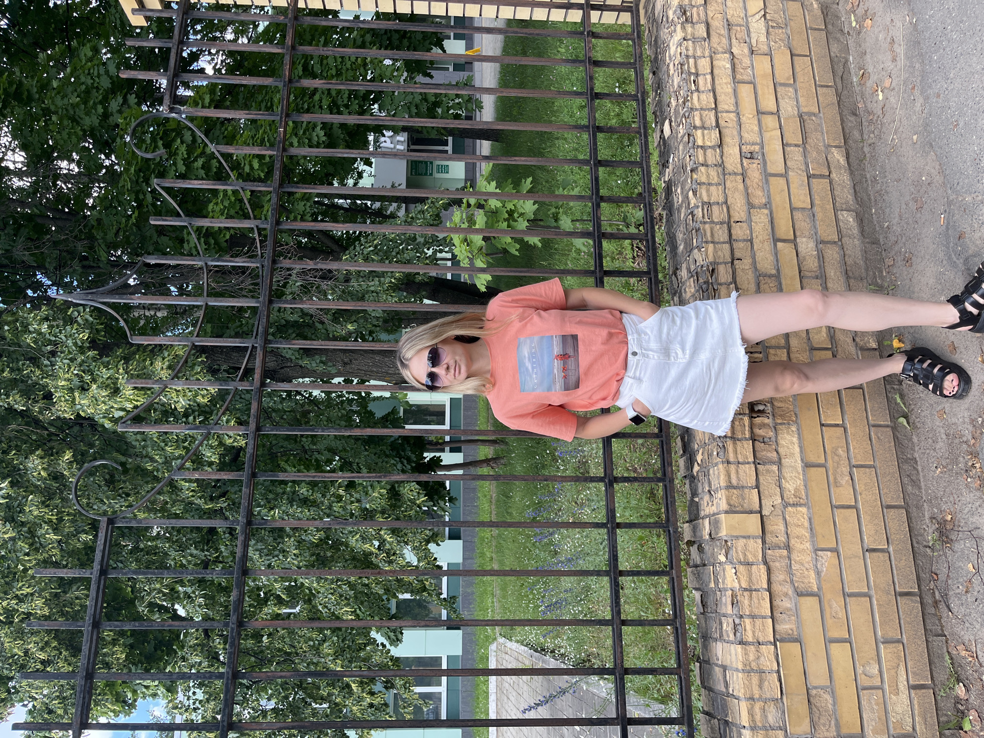 A blonde woman stands in front of a dark metal fence and a brick wall. She is wearing a coral t-shirt with a picture on it, a white denim skirt, and black sandals. She has sunglasses on and her hands are in her pockets. Behind her is a green lawn with scattered wildflowers, trees, and a building with windows. The scene is outdoors in a city, likely Chaikovsky, Russia.