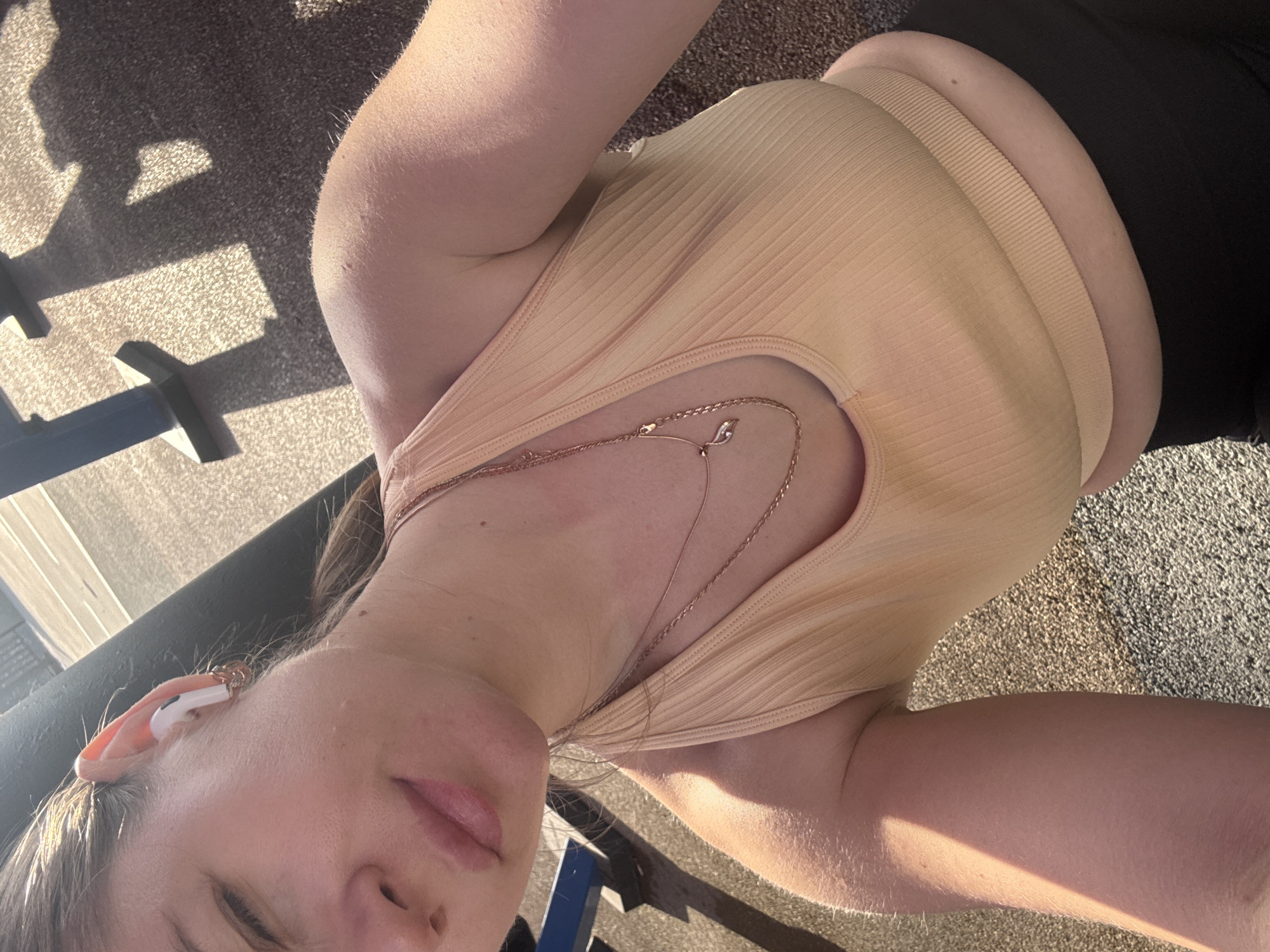 The image is a selfie of a woman wearing a light orange ribbed tank top and black pants, possibly leggings. She is wearing a pair of white wireless earbuds in her left ear and two delicate gold necklaces. The background shows a rough asphalt surface and part of a blue and grey structure, possibly a park bench or playground equipment. The lighting suggests it's daytime, with harsh shadows cast on the ground. Without additional information, pinpointing the location within Batumi, Georgia, is not possible.