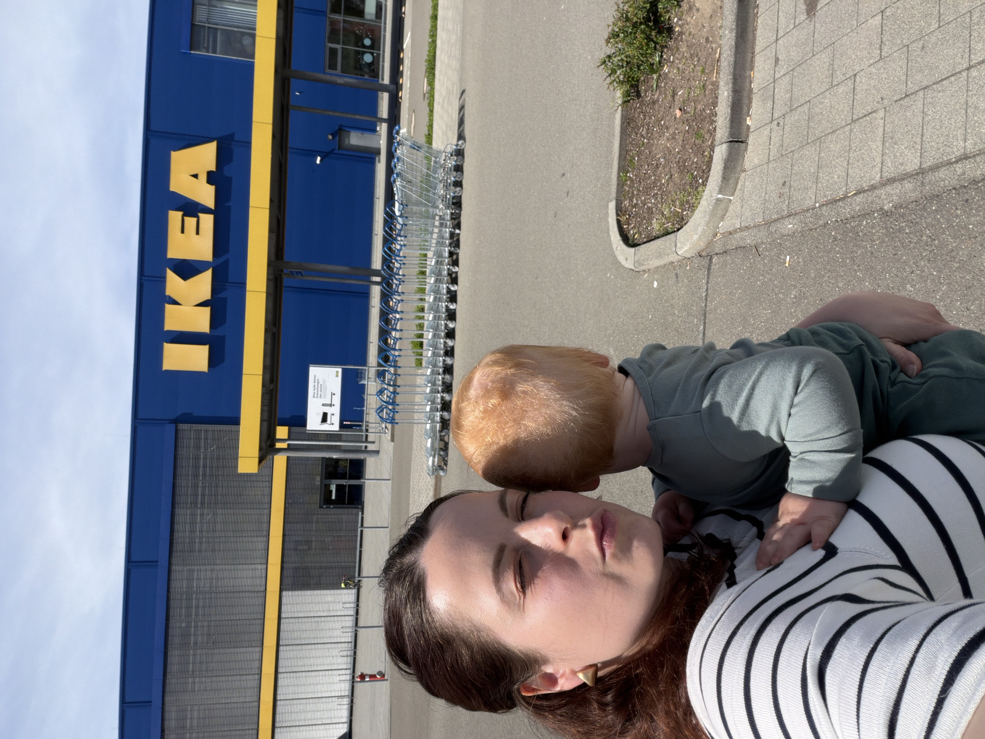 The image captures a selfie of a woman and a child outside an IKEA store on a bright, sunny day in Kaiserslautern, Germany. The woman, with long dark hair, is positioned in the foreground, looking towards the camera with her eyes partially closed, possibly squinting from the sun. She is wearing a white and black striped top. A young child, with light reddish-blonde hair and wearing a solid green long-sleeved outfit, is leaning against her shoulder, facing away from the camera.
In the background, to the left, is the distinct blue and yellow facade of an IKEA building, with the prominent yellow "IKEA" logo vertically displayed. A long row of silver shopping carts with blue handles is visible lined up next to the building. The scene takes place on a paved outdoor area, possibly a parking lot or pedestrian walkway. To the right of the shopping carts, there's a paved pathway and a small planted area with dirt and some greenery next to a curb. The sky is clear and light blue.
A white sign is visible near the shopping carts, with a graphic of a table and chair. The text on the sign reads: "Bring deine eigene Tragetasche oder Einkaufstasche mit!" (Bring your own carrying bag or shopping bag!) and "Schonen wir gemeinsam unsere Umwelt." (Let's protect our environment together.).