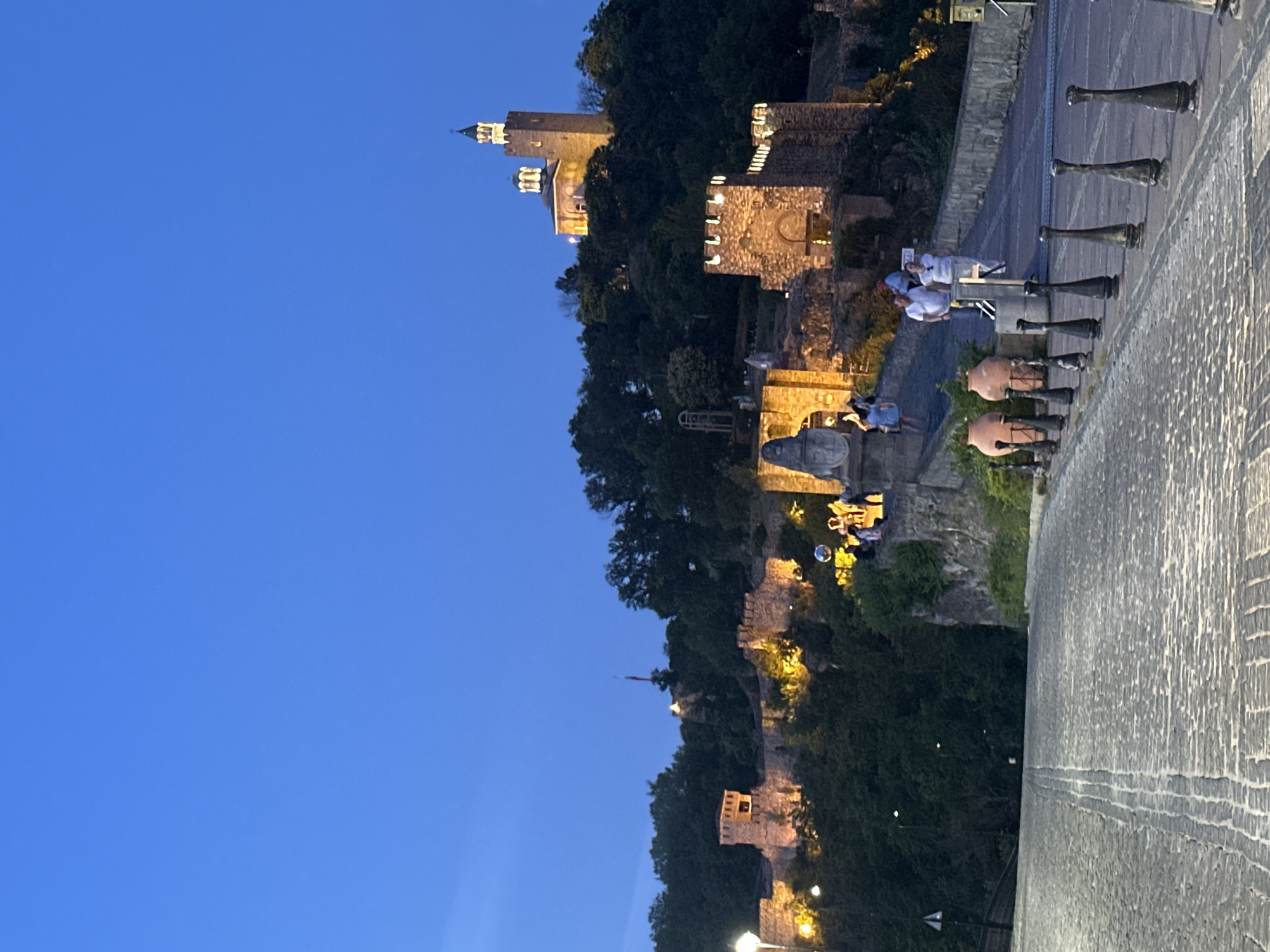 The image depicts a twilight scene at a historic fortified location, possibly a castle or citadel, in Veliko Tarnovo, Bulgaria. The sky is a deep blue, suggesting dusk or early evening. The architecture is characterized by stone walls, battlements, and towers, with some structures illuminated by warm, artificial lights, highlighting their textures and architectural details. Dense greenery, likely trees, flanks the fortifications, creating a stark contrast between the natural and man-made elements.
In the foreground, to the right, a paved walkway or parapet with a textured surface is visible, leading the eye towards the castle. Along this walkway, a row of dark, cannon-like objects are positioned at intervals. Further along, several people are seen. Three individuals are gathered near a barrier, appearing to observe the scenery or interact with each other. They are dressed in light-colored clothing. Another group of people is visible further down, also seemingly engaged in leisure activity. Two large terracotta pots are also present near the cannons.
The overall atmosphere is serene and somewhat majestic, conveying a sense of historical grandeur illuminated by the gentle evening light. The time of day is clearly marked by the blue sky and artificial lighting on the historical structures. No visible text is present in the image.