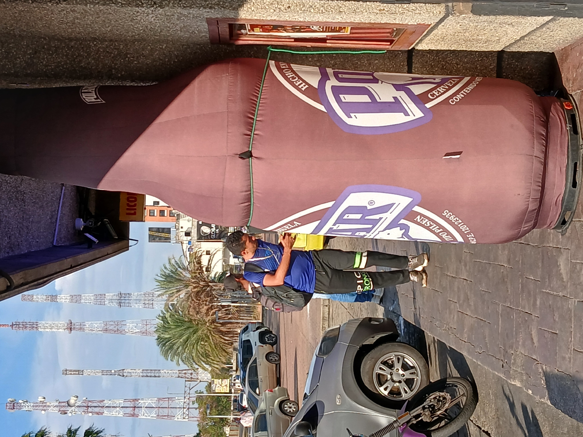 A giant inflatable beer bottle stands prominently on an urban street in Ciudad Guayana, Venezuela, under a clear, blue sky. The large, maroon-colored inflatable is shaped like a beer bottle, secured by green ropes along its sides. Its design features the brand name "POLAR" near the neck and a distinctive white and purple "P" logo on its body. Additional text visible on the bottle includes "HECHO EN", "CERVEZA", "CONTENIDO", "PILSEN", "NC.P.E. 120123935", and "AR".
A man wearing a blue t-shirt, black pants with green stripes (one leg showing "STRONG"), and a backpack stands near the base of the inflatable, looking down at a yellow object he holds. Another person is partially visible behind him. In the foreground, a silver hatchback car and part of a motorcycle wheel are visible, parked on a paved street next to a textured sidewalk.
The background reveals several tall communication towers against the bright sky, interspersed with green palm trees, indicating a warm, sunny climate. Other cars are parked further down the street. A sign on a nearby building displays "LICO", likely part of "LICOR" or "LICORERIA", and a distant sign shows "GLOBAL" and "QUELA". The scene depicts a sunny daytime setting, suggesting a promotional display or advertisement for the beer brand in a bustling urban environment.