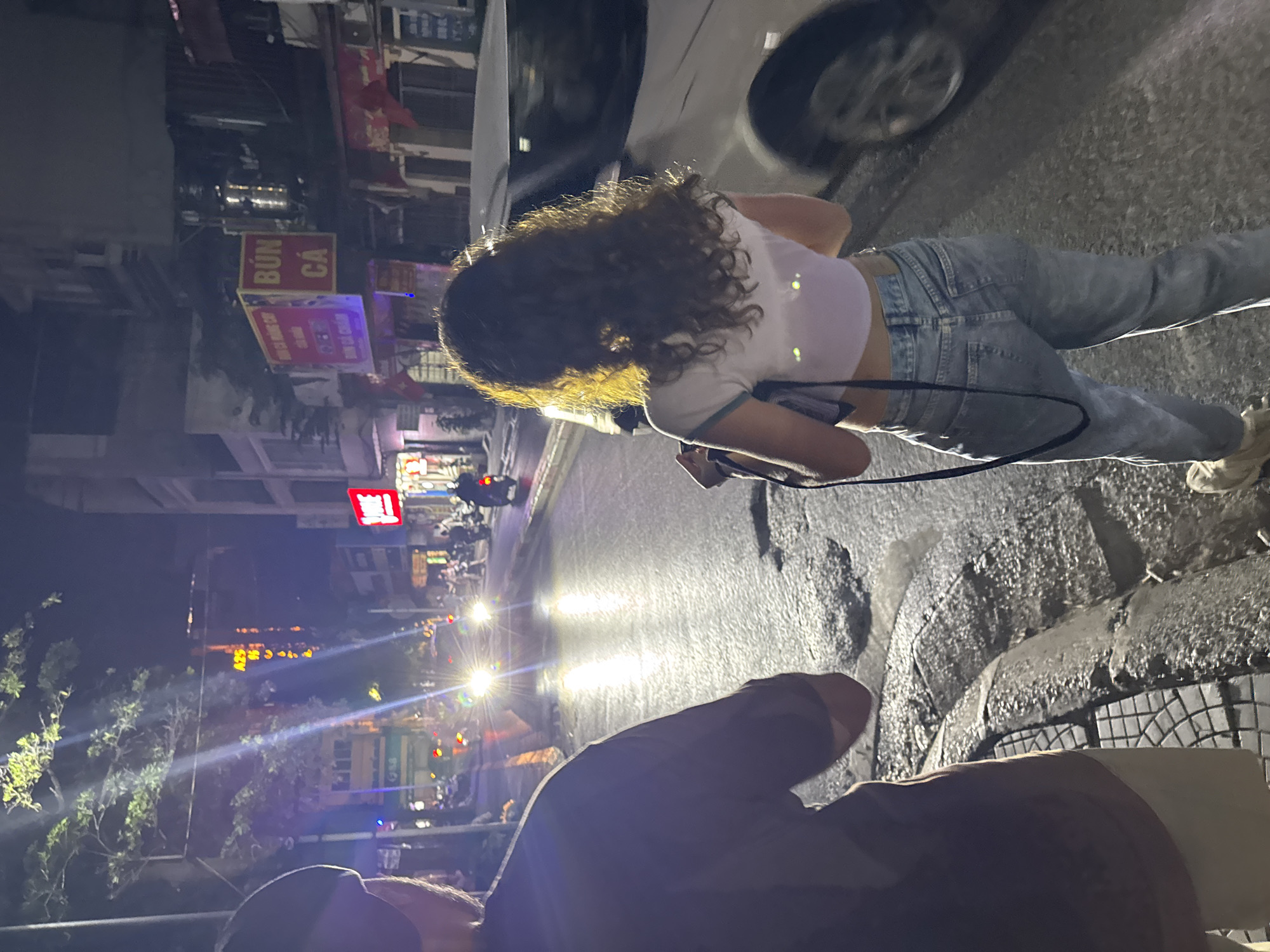 The image captures a street scene at night in Hanoi, Vietnam. In the foreground, a person with a dark shirt and light-colored shorts is partially visible on the left, facing away from the camera. To their right, a young woman with curly hair, wearing a light purple crop top and light blue jeans, is also seen from behind. She is holding a black bag or purse with a strap. The street is wet, reflecting the headlights of vehicles and streetlights, indicating recent rain or a damp surface. Potholes are visible in the asphalt.
In the background, several motorcycles are on the street, some with their headlights on, illuminating the road. Buildings line the street, displaying various signs, some illuminated with neon lights. One prominent sign in red and yellow reads "BÚN CÁ," which translates to "fish vermicelli," a popular Vietnamese dish. Another sign, partially obscured, appears to have Vietnamese text and a yellow glow. The overall atmosphere is dimly lit, characteristic of a city at night, with a sense of movement and activity from the passing vehicles. The perspective suggests the photograph was taken from a slightly elevated position or while standing near the curb.