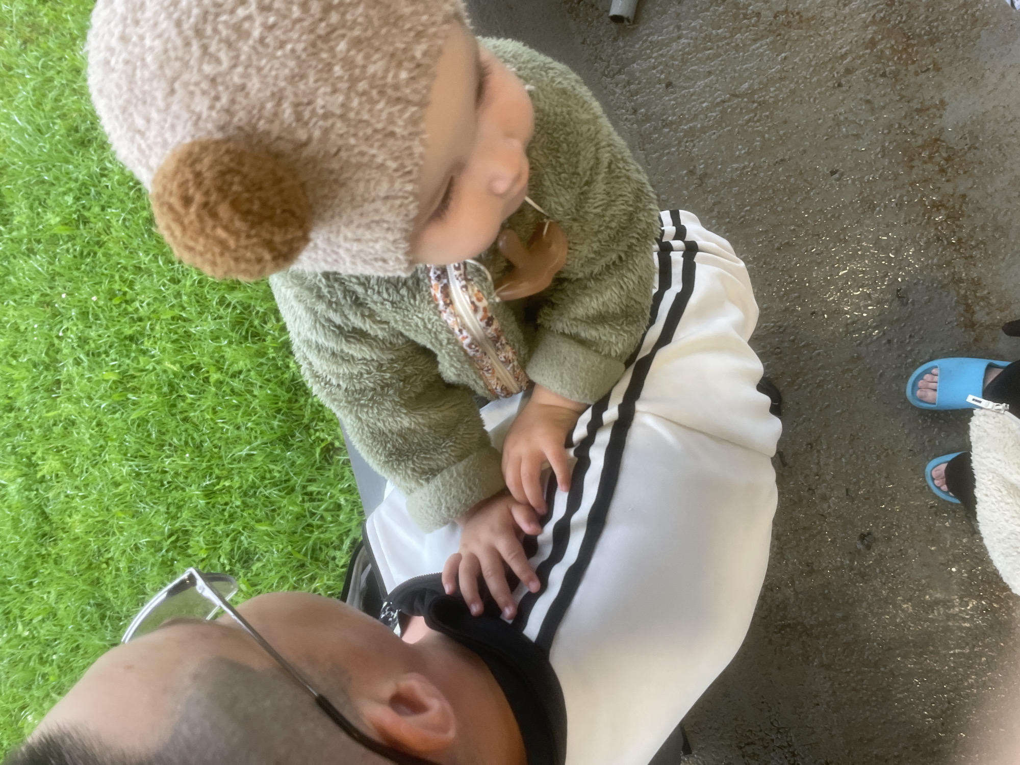 In Podshivalovo, Russia, a person wearing glasses and a black and white striped shirt is holding a baby dressed in a fuzzy green outfit and a bear-eared hat. The baby has a pacifier. They are outdoors on a wet, pebbly surface, with green grass in the background. Two pairs of feet are visible in the foreground, one wearing blue slides.