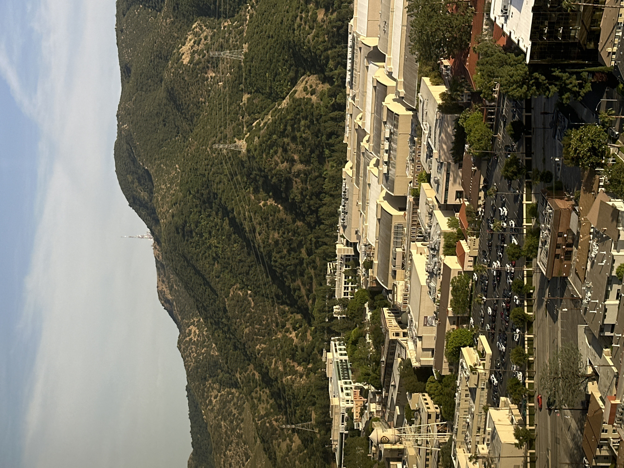 This high-angle photograph presents a dynamic view of an urban landscape nestled at the base of a significant mountain range, set in Burbank, United States of America.
On the left side, a vast, verdant mountain range, likely the Verdugo Mountains, dominates the scene. The mountains are densely covered with an array of trees and shrubs, showcasing deep green hues. Several prominent power lines, supported by tall metal pylons, traverse the rugged slopes, indicating essential infrastructure. On a distant peak, a slender communications tower or antenna stands tall against the sky. The sky above is a pale blue, adorned with sparse, wispy white clouds, suggesting clear weather.
The right side of the image features a bustling urban environment. Numerous multi-story buildings, predominantly in shades of beige, white, and grey, line the area. Many structures display modern architectural elements, including distinctive curved facades and balconies, typical of commercial or high-density residential complexes. A wide street, or a series of parallel roads, runs through the city, densely populated with vehicles. Many cars, appearing mostly white or dark-colored, are parked along the curbs and actively driving, indicating regular urban activity. Lush green trees are thoughtfully integrated into the streetscape, planted along sidewalks and between buildings, adding natural elements to the concrete jungle.
The image captures the scene during daylight hours under sunny conditions, evidenced by the bright illumination and distinct shadows cast by buildings and mountains. No individual people are clearly discernible, and no text is visible on any signs or structures. The photograph conveys a sense of vibrant daily life in a scenic urban setting, beautifully contrasted by the imposing natural grandeur of the mountains.