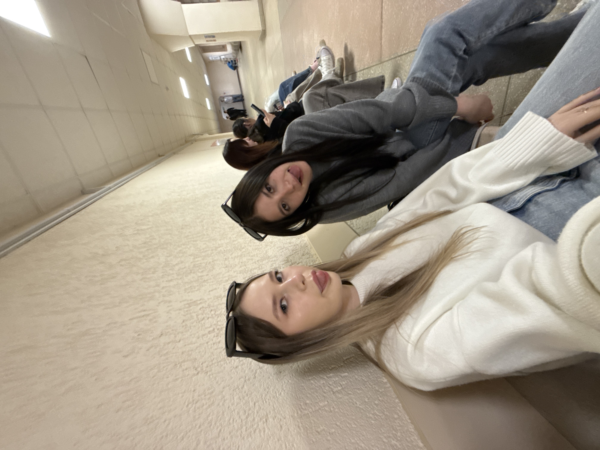 In a hallway in the city of Chaikovsky, Russia, a group of women sit along a wall, possibly waiting. Two women are prominent in the foreground. The woman closest to the camera has light brown hair and is wearing a white sweater and sunglasses. The second woman has long, dark hair, a grey sweater, and is also wearing sunglasses. Both women are sticking out their tongues. Several other women are visible in the background, also seated along the wall, some using phones. The hallway is long, with light walls and recessed lighting in the ceiling.