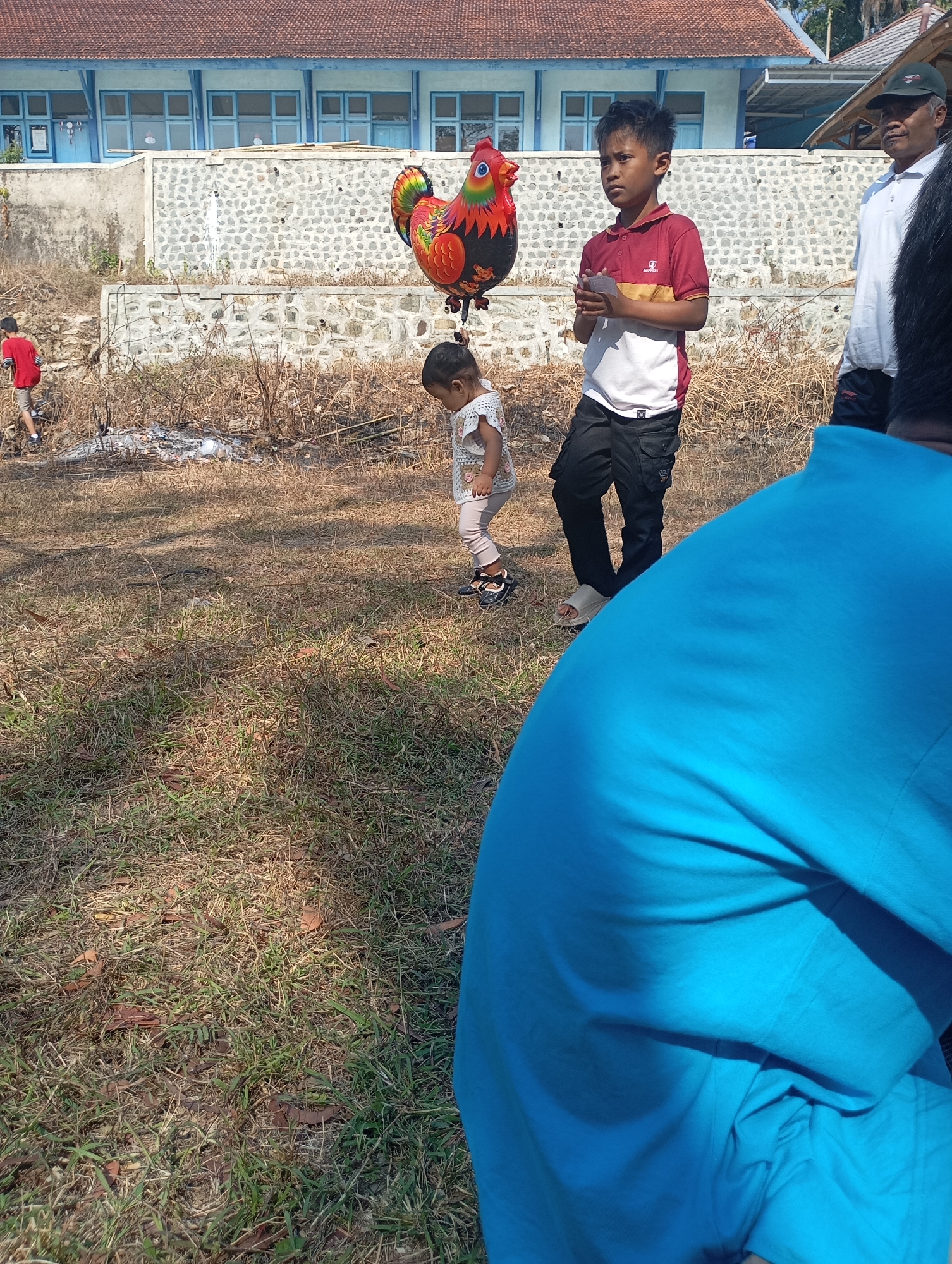 In Ketepung, Indonesia, on a dry, grassy field, a group of people are gathered near a stone wall and a building with a red-tiled roof. A young boy in a red and white polo shirt and black cargo pants holds a rooster-shaped balloon. Beside him, a young girl in a white dress stands, her gaze directed downwards. Further back, a man in a white polo shirt and a cap watches the scene. In the foreground, a person wearing a bright blue shirt is partially visible, with their back to the camera. The environment is rustic, with dry brush and scattered leaves on the ground. A person in a red shirt is visible in the background, near some discarded debris.