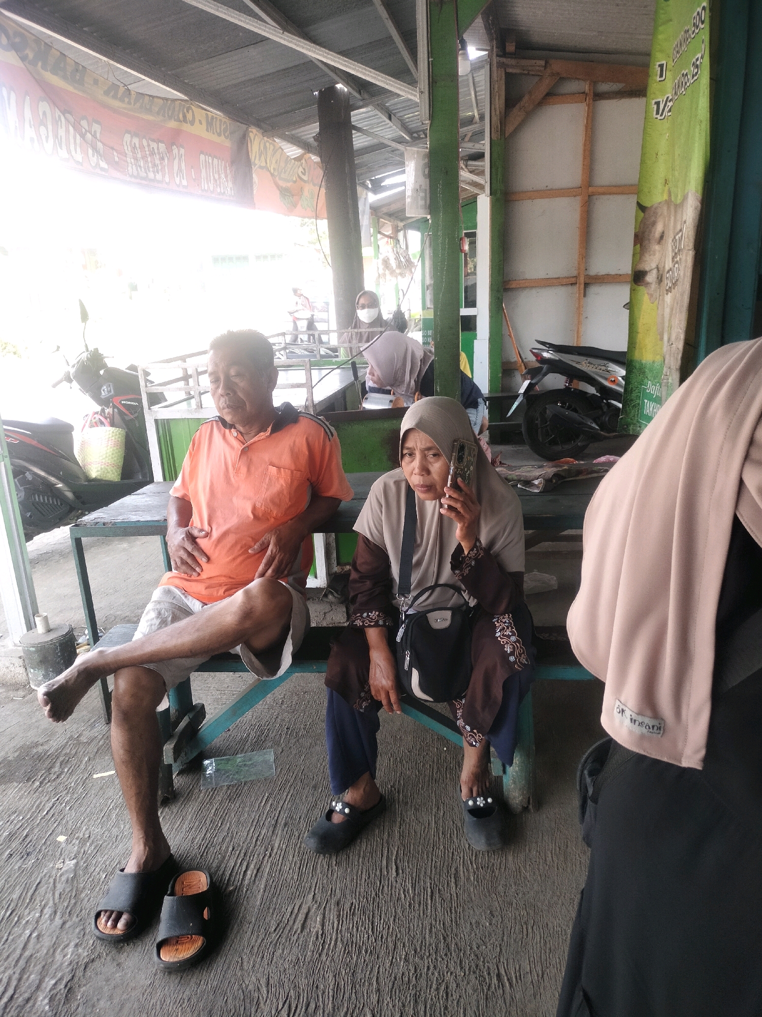 The image depicts an outdoor scene at what appears to be a roadside food stall in Tersono, Indonesia. Two individuals are seated on a bench in front of a table. On the left, an older man wearing an orange polo shirt and grey shorts is seated with his hands on his stomach and one leg crossed over the other. To his right, a woman in a beige hijab and a brown and beige patterned dress is holding a mobile phone to her ear and looking towards the camera. Her purse is slung over her shoulder. The ground is a textured concrete surface. Behind the individuals, a green metal table and bench are visible. In the background, a motorcycle is parked, and a stall structure with a corrugated metal roof and wooden supports is present. A large banner with Indonesian text and an image of a cow is hanging on the right side of the frame. Another banner with text is visible on the left. The overall environment suggests a casual, possibly commercial, setting in a town or village.