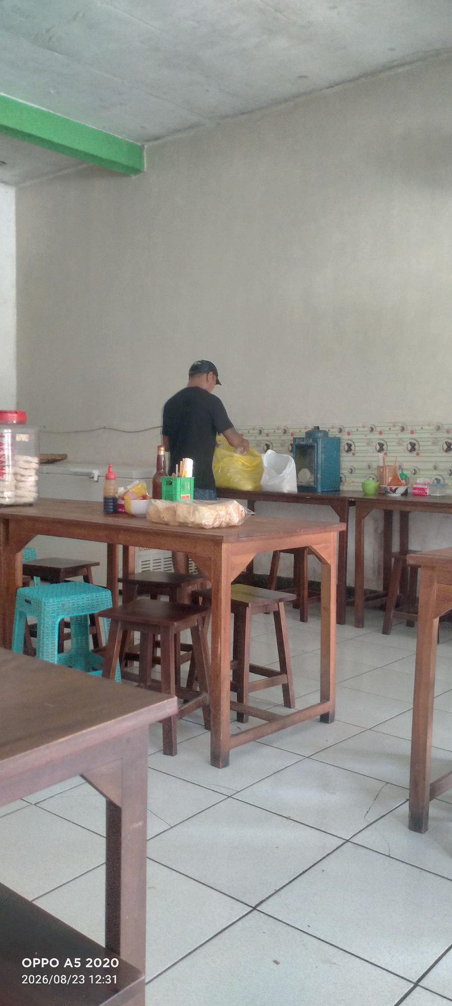 The image depicts the interior of a simple eatery, likely a warung, located in Dusun Satu, Indonesia. The walls are a neutral, light color, and the ceiling appears to be concrete with some green trim. The floor is tiled in a grid pattern.
In the center of the frame, a person wearing a dark t-shirt and a baseball cap is standing behind a wooden counter, sorting through a large yellow plastic bag. The counter is laden with various items, including bottles of condiments, a green plastic container with utensils, a bowl, and a package of what appears to be crackers or snacks. To the left of the counter, a clear plastic container filled with similar snack items is visible. In the background, another wooden counter with a blue appliance on it is present.
Several wooden tables and stools, as well as a bright teal plastic stool, are arranged in the dining area. The overall impression is one of a functional, unpretentious establishment. The timestamp on the image indicates it was taken on August 23, 2026, at 12:31.