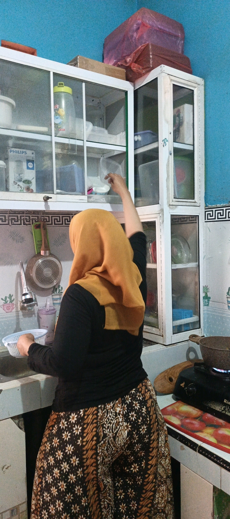In the city of Pandes, Indonesia, a woman wearing a yellow hijab and a black long-sleeved shirt is standing in a kitchen. She is facing away from the camera, with her right arm extended, reaching into a cabinet. Her pants feature a batik pattern of white flowers on a black background, with brown stripes. She is holding a white basket in her left hand, which rests on the edge of a sink. To her left, a faucet and a drain are visible. Above the sink, a cabinet with glass doors contains various kitchen items, including containers and a bottle with a yellow lid. Behind the woman, another cabinet is partially open, revealing more items. In the foreground, a small gas stove is on, with a pot on top. A cutting board is placed next to the stove. The kitchen walls are painted blue, and the cabinets are white with some rust.
