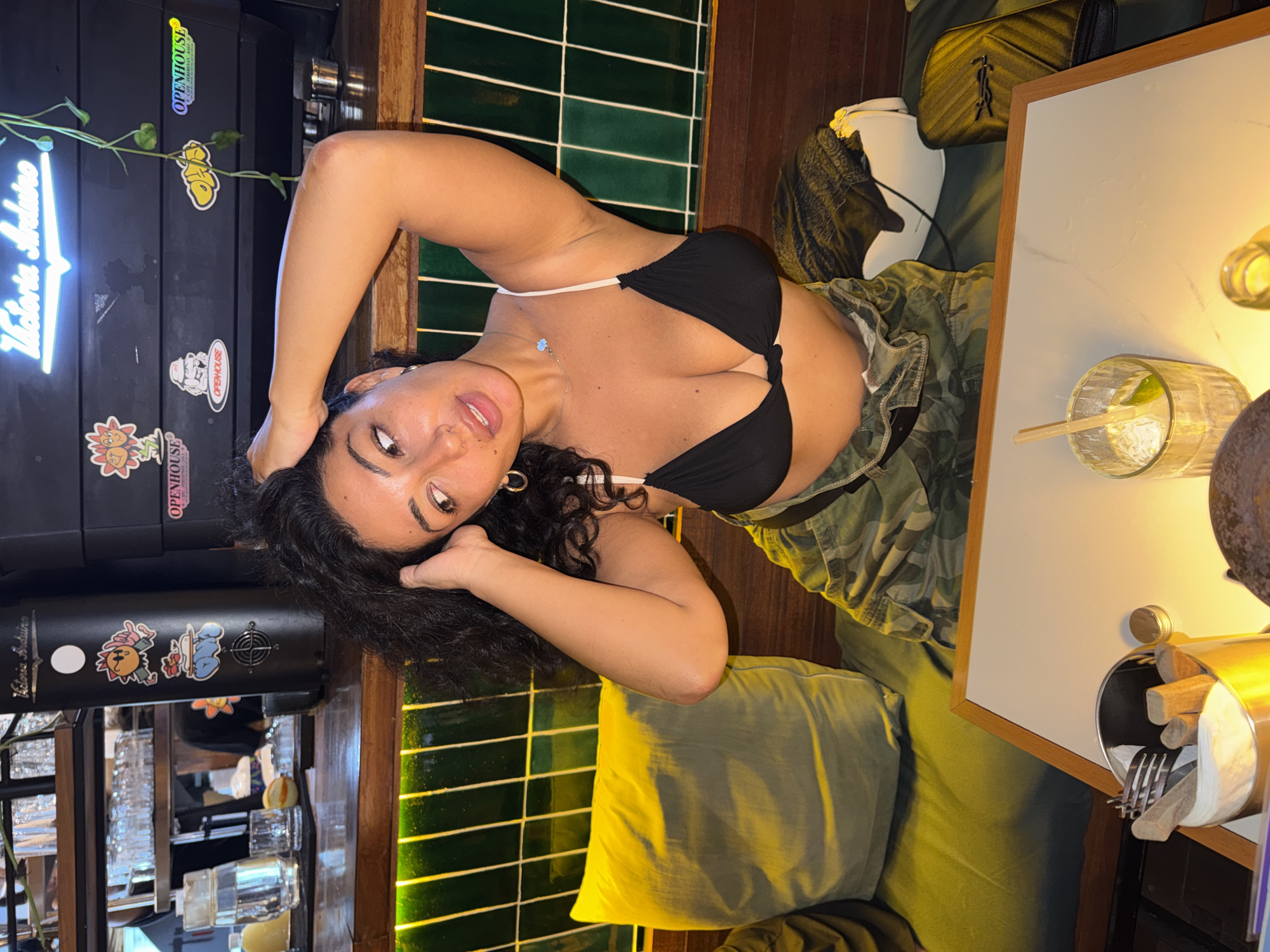 A woman with curly dark hair is seated at a table in a dimly lit establishment, possibly a cafe or bar, with a wooden counter and green tiled backsplash. She is wearing a black bikini top and camouflage pants. Her right arm is raised, with her hand in her hair, and her head is tilted to the side. A glass of ice and lime, along with a metal utensil holder, are on the table in front of her. The background includes a coffee machine adorned with stickers, shelves stocked with glasses, and plants hanging from the ceiling. The location appears to be in Mengwi, Indonesia, based on sticker text referencing "OPENHOUSE CAFE PERANAKAN BALI".