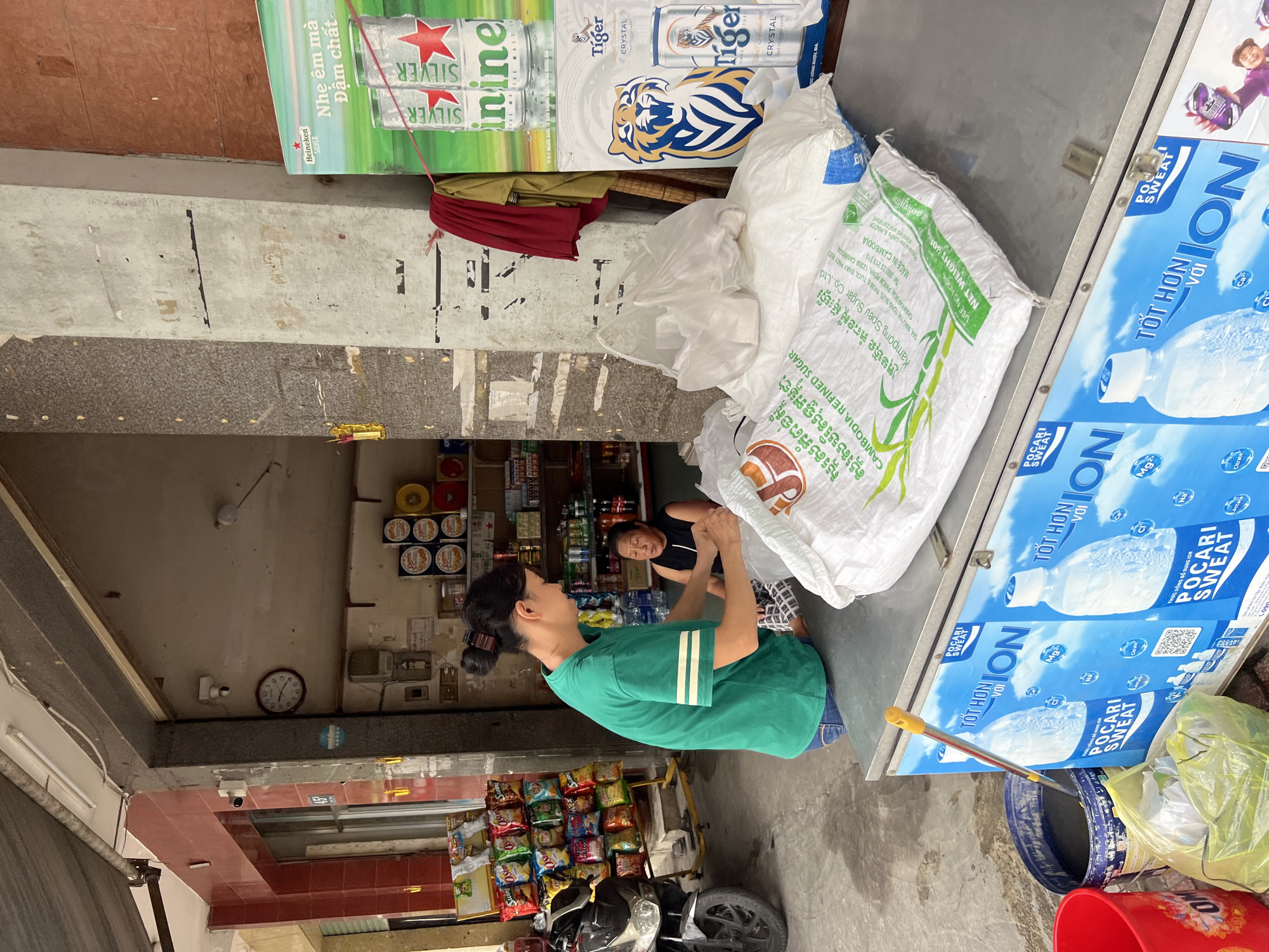 The image depicts an outdoor scene in Nha Trang, Vietnam, likely in front of a small convenience store or market. In the foreground, a person wearing a green t-shirt and blue jeans is leaning over a metal counter, handling a large white bag of sugar that has text in Khmer and English, indicating it is "CAMBODIA REFINED SUGAR" from "Kampong Speu Sugar Co., Ltd." Next to the sugar bag are several plastic bags and a large blue bucket containing liquid, with a yellow-handled broom partially submerged. Behind the person in green, a second person, an older woman wearing a black tank top, is seated and appears to be talking or gesturing. The counter is an open-fronted freezer, decorated with posters of "POCARI SWEAT" drinks. In the background, shelves are stocked with various goods, including snacks and bottles of beverages. A motorcycle is parked to the left. The environment appears to be a street scene with a covered walkway and building facades. A large poster advertising Heineken Silver beer and Tiger Crystal beer is visible on a pillar to the right.