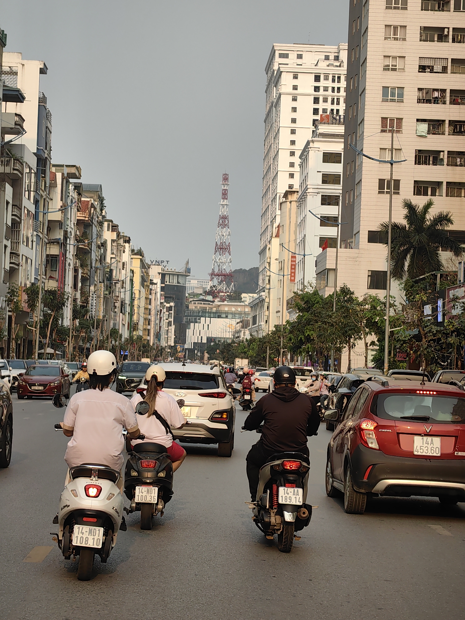 The image captures a bustling street scene in Hong Gai Ward, Vietnam, filled with vehicular traffic. A tall, red and white lattice communications tower rises in the distance, between buildings. Dominating the foreground are three motorbikes navigating the crowded street, each ridden by a single person. The riders are wearing helmets. Two of the motorcycles are relatively close together. A red car is visible to the right. The street is lined with multi-story buildings of varying architectural styles. The environment appears to be urban and somewhat densely populated. License plate details can be made out. The sky is overcast, suggesting a cloudy day.