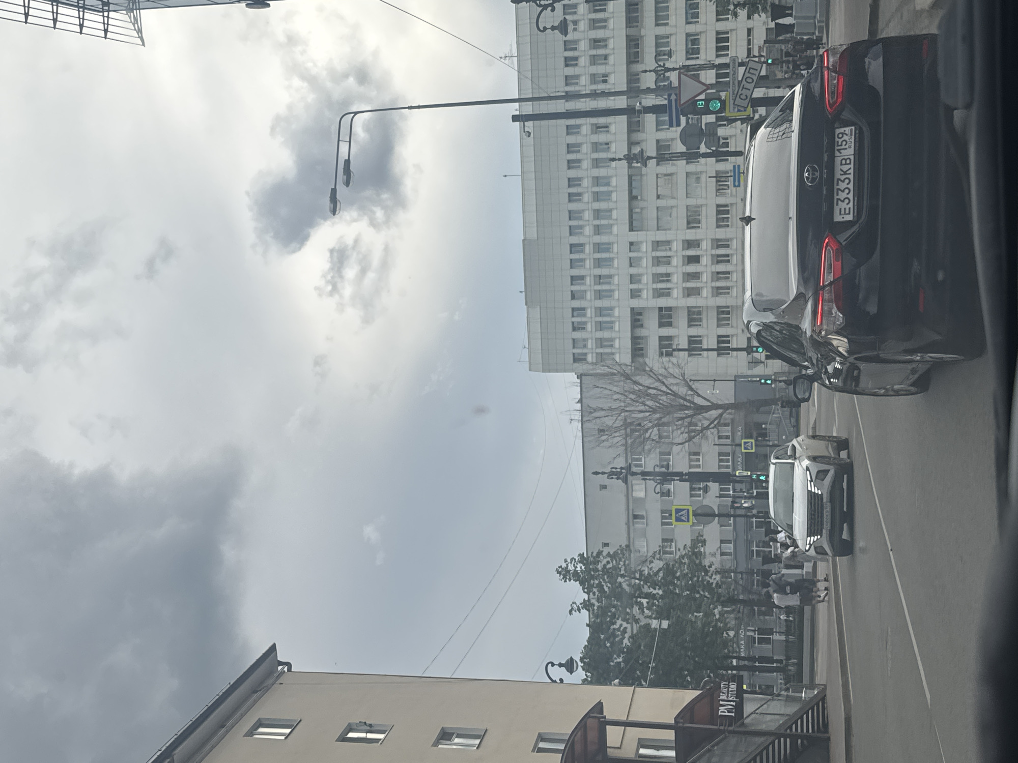 The image captures a street scene in the city of Perm, Russia, under an overcast sky with dramatic clouds. In the foreground, the rear of a black Toyota Camry is prominently displayed, showing its license plate which reads "E333KB 159 RUS". The car is in motion, heading away from the viewer. To the left of the Camry, a white SUV, possibly a Hyundai Tucson, is also in motion, approaching the viewer.
The environment features a wide city street with multiple lanes. On the left side of the street, a light-colored, multi-story building with visible windows is present, with a covered entrance and stairs leading up. Further down the street, across the road, a large, long, light-colored administrative building with numerous windows dominates the background. Between these buildings, trees and streetlights are visible.
Traffic signals and street signs are scattered throughout the scene. A triangular yield sign and a rectangular sign with the Russian word "СТОП" (STOP) are clearly visible. Several pedestrian crossings are also indicated by road markings.
A few individuals are seen on the sidewalk in the mid-ground, walking away from the viewer. The overall impression is that of a typical urban street in Russia, with a mix of modern and older architecture and active vehicular and pedestrian traffic.