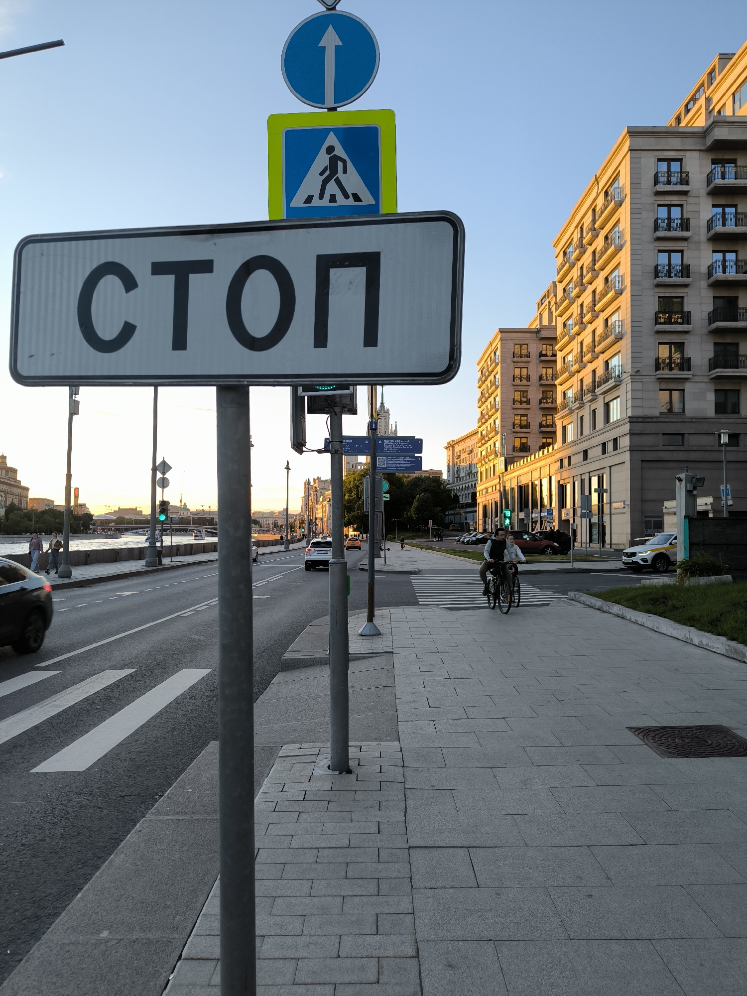 This image captures a street scene in Moscow, Russia, near the Moskva River. A prominent "STOP" sign in Russian is in the foreground, along with traffic lights and pedestrian crossing signs. To the right, a modern building with multiple balconies stands tall. A couple is seen cycling on the sidewalk. Cars are present on the road, and a bridge is visible in the distance, spanning the river. The setting appears to be during the golden hour, with the sun casting a warm glow on the buildings.