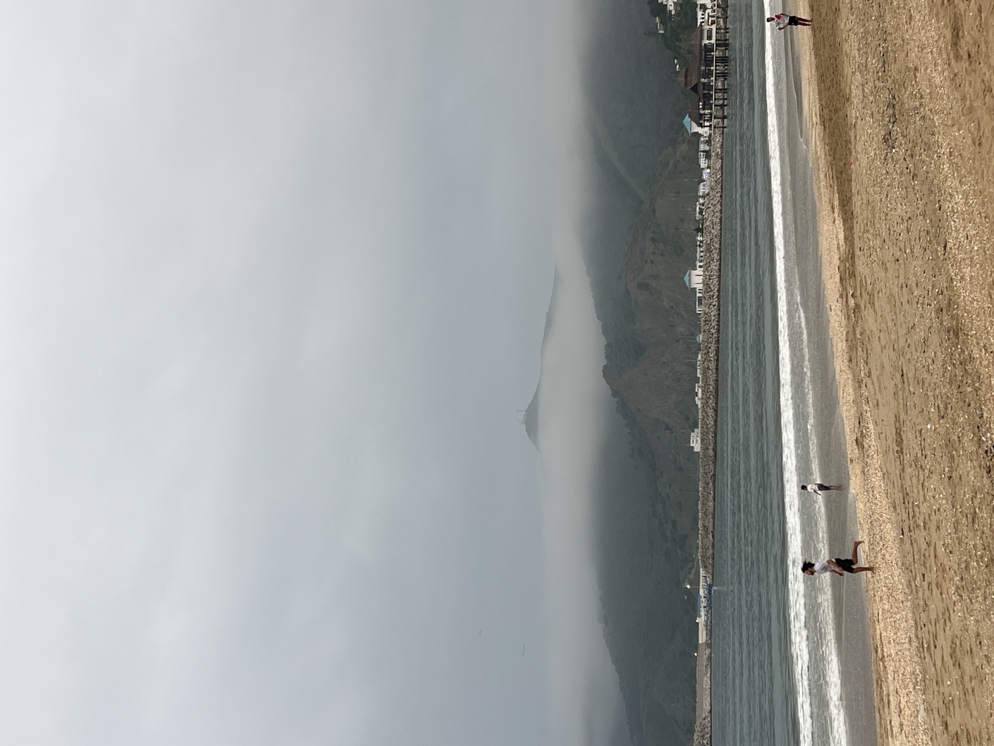The image displays a coastal scene in M'Diq, Morocco, under heavily overcast and foggy conditions. The left portion of the image shows a vast expanse of grey sky and dense mist, which obscures the upper parts of a mountain range. The base of the mountains reveals a developed area with numerous buildings, some featuring blue roofs, situated behind a long, grey seawall or breakwater.
To the right, the sea meets a sandy beach. The water shows gentle, white-foamed waves breaking along the shore. The beach itself is light brown, covered with sand and small pebbles or shells. Several people are visible on the beach and at the water's edge. One individual stands in the shallow surf, wearing dark shorts and a light top. Closer to the foreground, another person in similar attire appears to be bent over or in motion near the waterline. Further along the beach, another figure is standing near the water.
The thick fog creates an atmospheric and somewhat muted quality to the landscape, diminishing visibility into the distance and lending a serene, misty ambiance to the scene. No discernible text is visible in the image.