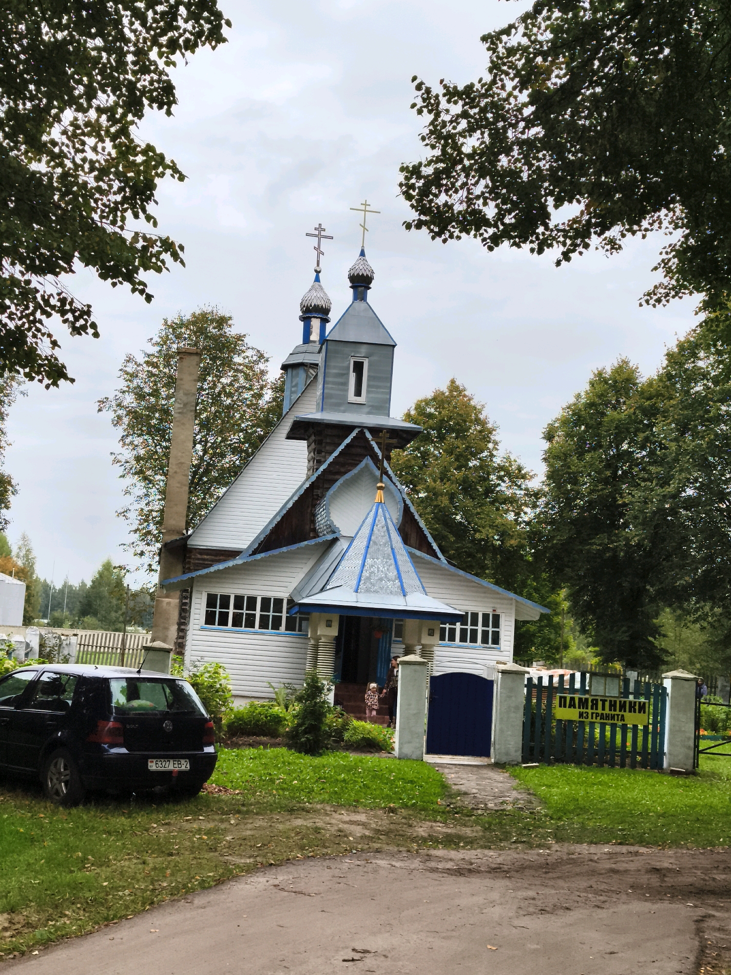 The image shows a white wooden church with blue trim and a blue roof. The church has two onion domes topped with crosses. In front of the church is a blue gate with a yellow sign that says "ПАМЯТНИКИ ИЗ ГРАНИТА" in Cyrillic, which translates to "MONUMENTS FROM GRANITE." To the left of the church is a black Volkswagen Golf with a Belarusian license plate. The church is surrounded by trees and grass, and there is a dirt road in the foreground. Two people, a woman and a child, are visible near the entrance of the church. The scene is overcast.