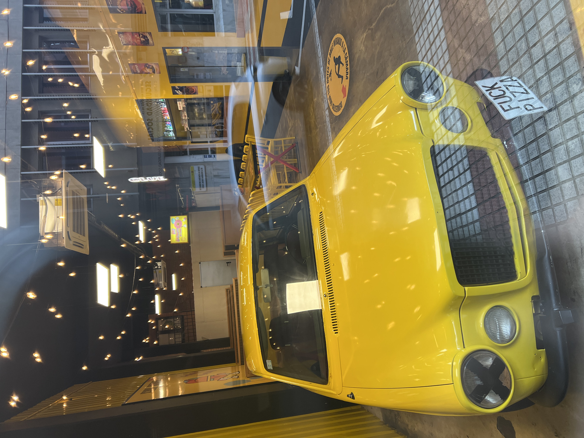 The image captures an indoor scene, likely a themed restaurant or entertainment venue in Las Palmas, Spain, viewed through a reflective glass surface. The central subject is a vibrant yellow car, possibly a custom build or classic model, prominently displayed. It features distinct round headlights, one of which is marked with an 'X' using black tape, and a black front grille. A novelty license plate, visible upside down, reads "FUCK PIZZA".
The setting includes a concrete floor with yellow and black striped markings, reminiscent of a road or parking lot. A circular floor decal is visible, featuring a stylized figure and the Spanish text "DEMUESTRA TUS PASOS DE BAILE MIRANDO A LA CÁMARA" (Demonstrate your dance moves looking at the camera) and "CLUB". In the background, bright yellow tables and chairs suggest a dining or seating area.
The glass through which the photo is taken creates strong reflections, revealing numerous string lights, rectangular light fixtures, and other commercial signage. Words like "DONUTS" and "Restro" are discernible in the reflections, along with parts of posters and banners. The abundance of artificial lighting and reflections suggests an evening or nighttime setting. The overall atmosphere appears quirky and playful, centered around the distinctive yellow vehicle. No people are visible.