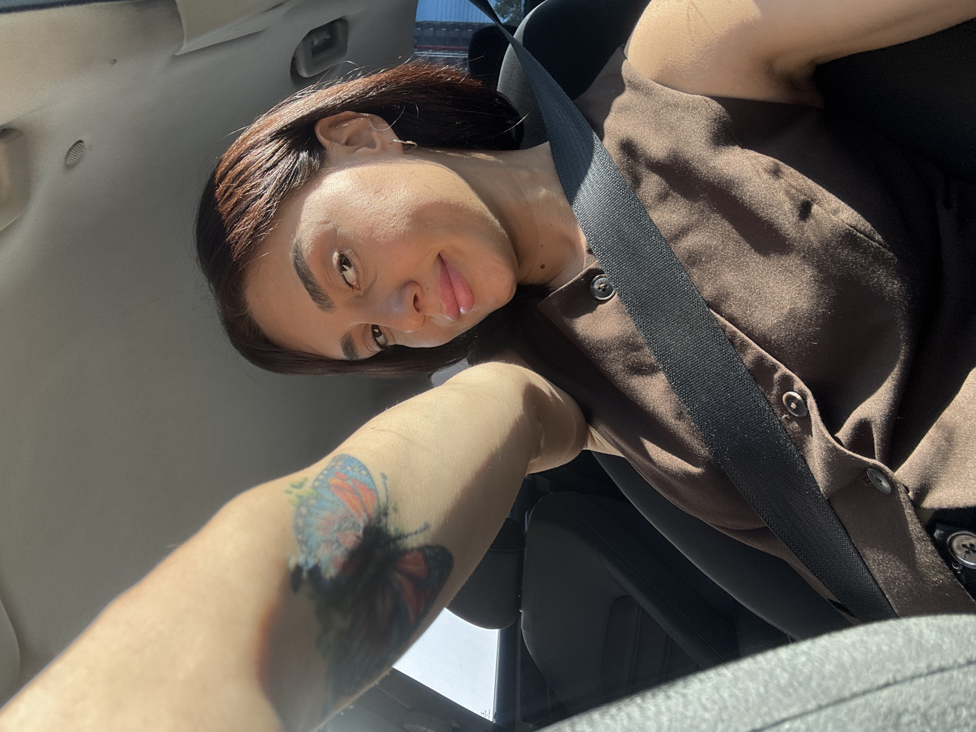 The image shows a woman with shoulder-length brown hair and fair skin, wearing a brown sleeveless top and a seatbelt. She has a butterfly tattoo on her left arm. She is sitting in the driver's seat of a car, with her arm extended as if taking a selfie. The interior of the car, including the seats and the headliner, is visible. The lighting suggests it is daytime, with sunlight streaming into the vehicle. The location is identified as Tbilisi, Georgia.