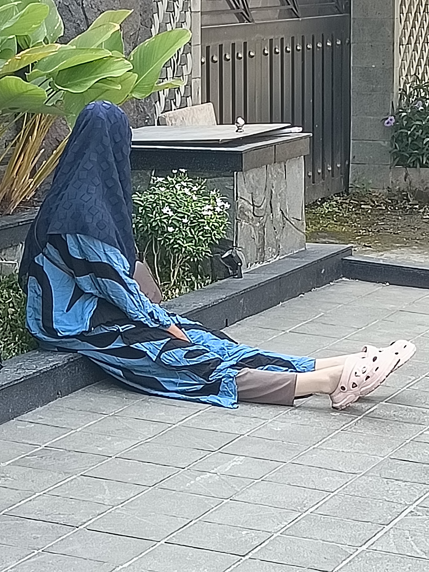A person wearing a dark blue patterned headscarf and a blue and black patterned long-sleeved tunic and brown pants is sitting on a tiled floor next to a raised planter box. The person is wearing light pink clogs with decorative holes. Behind the person is a dark stone wall and a metal fence. To the left of the person is a large leafy plant. To the right of the person, a small bush with white flowers is visible. The scene appears to be outdoors in a residential or public space, potentially in the city of Kediri, Indonesia, given the architectural style and vegetation.