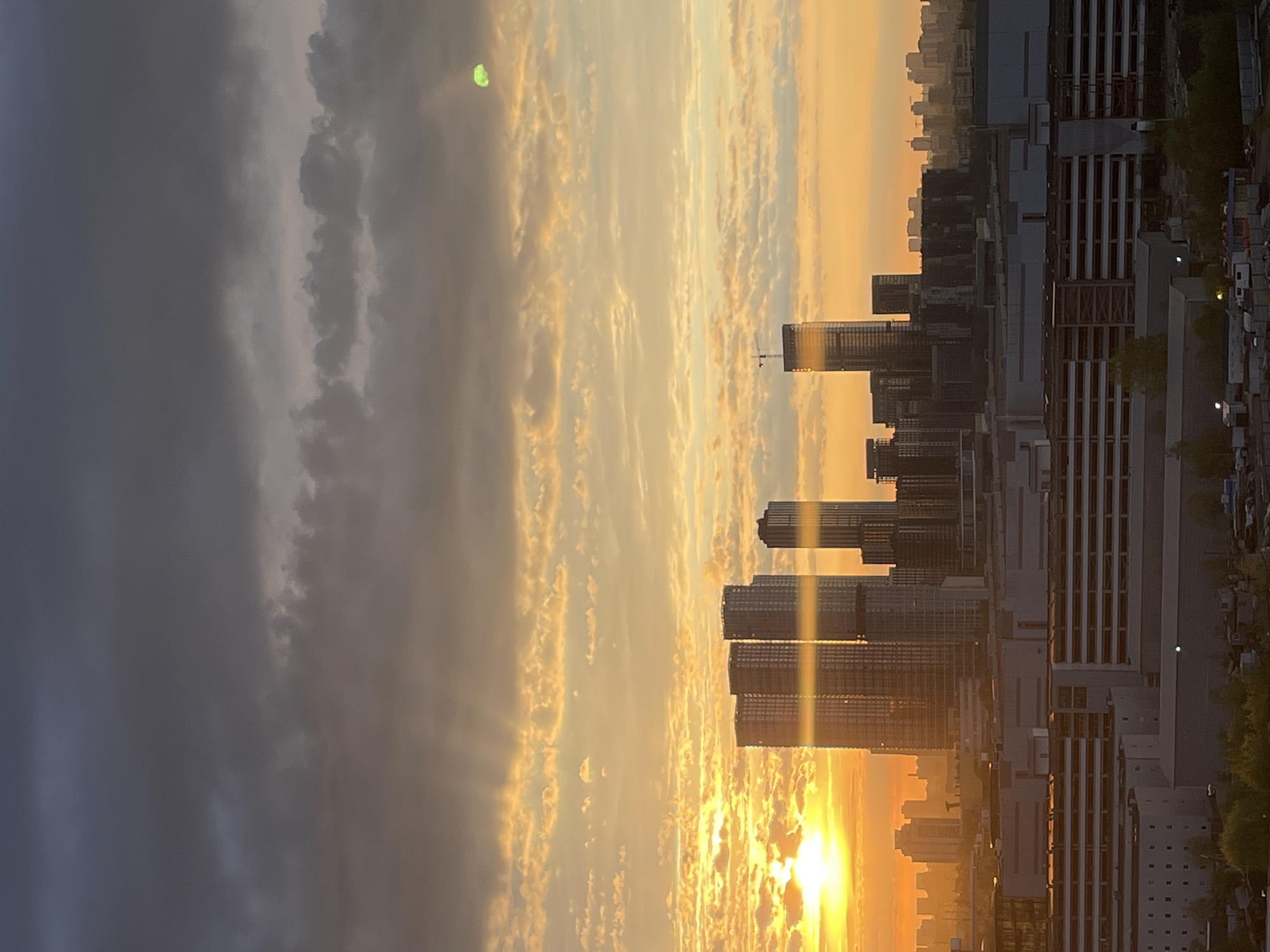 Here is a concise description of the image:
The image depicts a sunset over a city skyline. The sun is low on the horizon, casting a golden glow over the clouds and reflecting off the buildings. There are multiple high-rise buildings in the frame, some under construction. The left side of the image shows dark storm clouds moving in. The location is a city with a mix of completed buildings and ongoing construction.