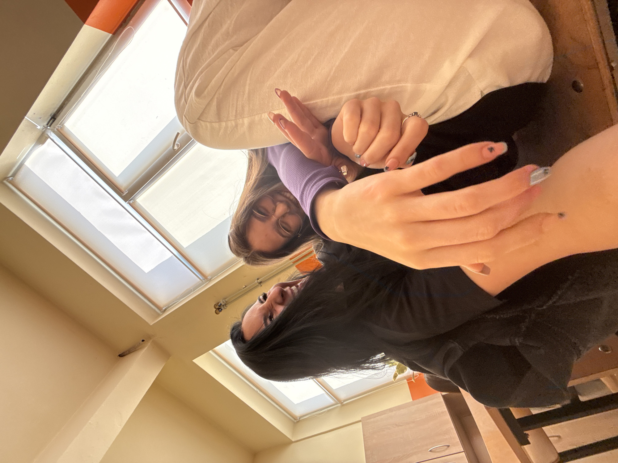 The image shows two people in a room with two skylights. One person is wearing a purple top and glasses, and is positioned towards the upper left of the frame. The other person, wearing a black top, is positioned towards the lower right. Both people are facing the camera. The room has light-colored walls and ceiling. A piece of furniture, possibly a dresser, is visible in the lower right corner. The specific location within Sofia, Bulgaria cannot be determined from the image.
