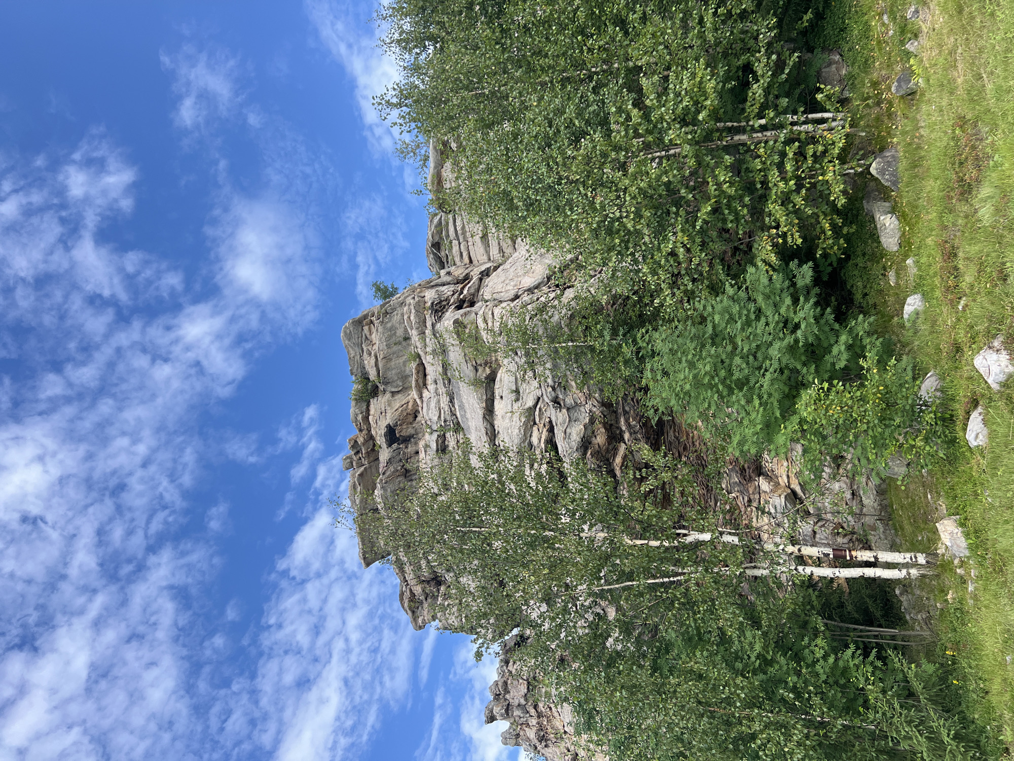 The image depicts a natural scene featuring a large rock formation against a backdrop of a blue sky with wispy white clouds. In the foreground, lush green trees and grass cover the ground. The rock formation is rugged and appears to be composed of layered stone. Several trees, including birch trees with white bark, are positioned in front of the rocks. The overall environment suggests an outdoor, mountainous or hilly area. This scene is located in the vicinity of the Ural Mountains, a region that includes the city of Gubakha, Russia, known for its natural landscapes and rock formations.