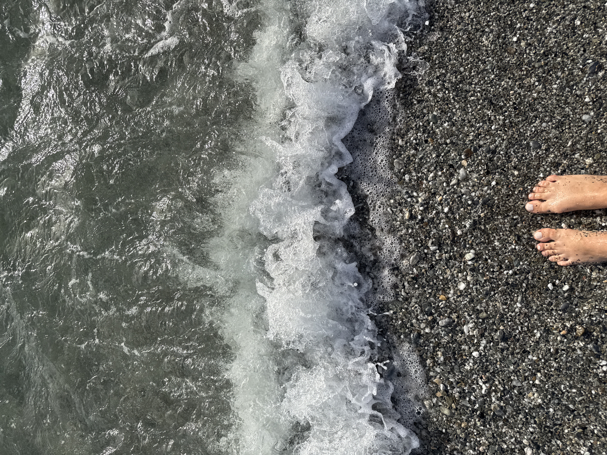 The image shows a top-down view of a beach with waves washing ashore. Two bare feet are visible on the right side of the image, standing on dark, pebbly sand. The water is dark gray and clear, with white foam from the waves. The scene appears to be a natural beach environment. Based on the provided information, the location is a beach in Lamezia Terme, Italy.