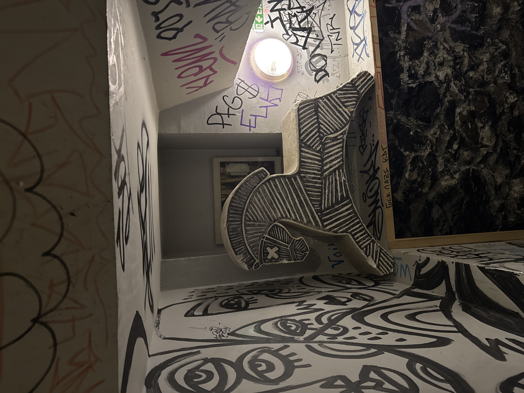 The scene depicts an interior view of the Hadji Dimitar underpass in Sofia, Bulgaria, characterized by concrete walls and ceiling extensively covered in graffiti. Centrally positioned is a profile silhouette of a horse, approximately 60-70 cm tall, constructed from a light-colored material with dark, linear, geometric patterns etched or painted across its body, head, and mane. An "X" mark covers its eye. The horse rests on a narrow ledge or beam.
The wall to the left of the horse is predominantly white, covered with intricate black graffiti featuring numerous stylized eyes and abstract curvilinear designs. The wall and ceiling section to the right of the horse are densely packed with overlapping graffiti tags in various colors, including black, pink, purple, white, red, and blue. Visible tags include "KLOW," "PFS," "ELVS," "DOVA," and "TNR." A phrase "FUCK NAZI KAT" is stenciled in red on a horizontal beam beneath the horse, and "Voilà" is written in blue below the horse itself.
A single circular, recessed light fixture with a bright, cool-toned bulb illuminates the right side of the scene, casting a faint purple glow on the surrounding graffiti. A green emergency exit sign icon is partially visible to the immediate right of the light. Behind the horse, in a recessed alcove, a framed object, possibly a mirror or picture, is partially visible. The lower section of the wall below the horse features a dark, heavily textured, possibly stone or stucco surface. No people, discernible actions, or interactions are visible.
