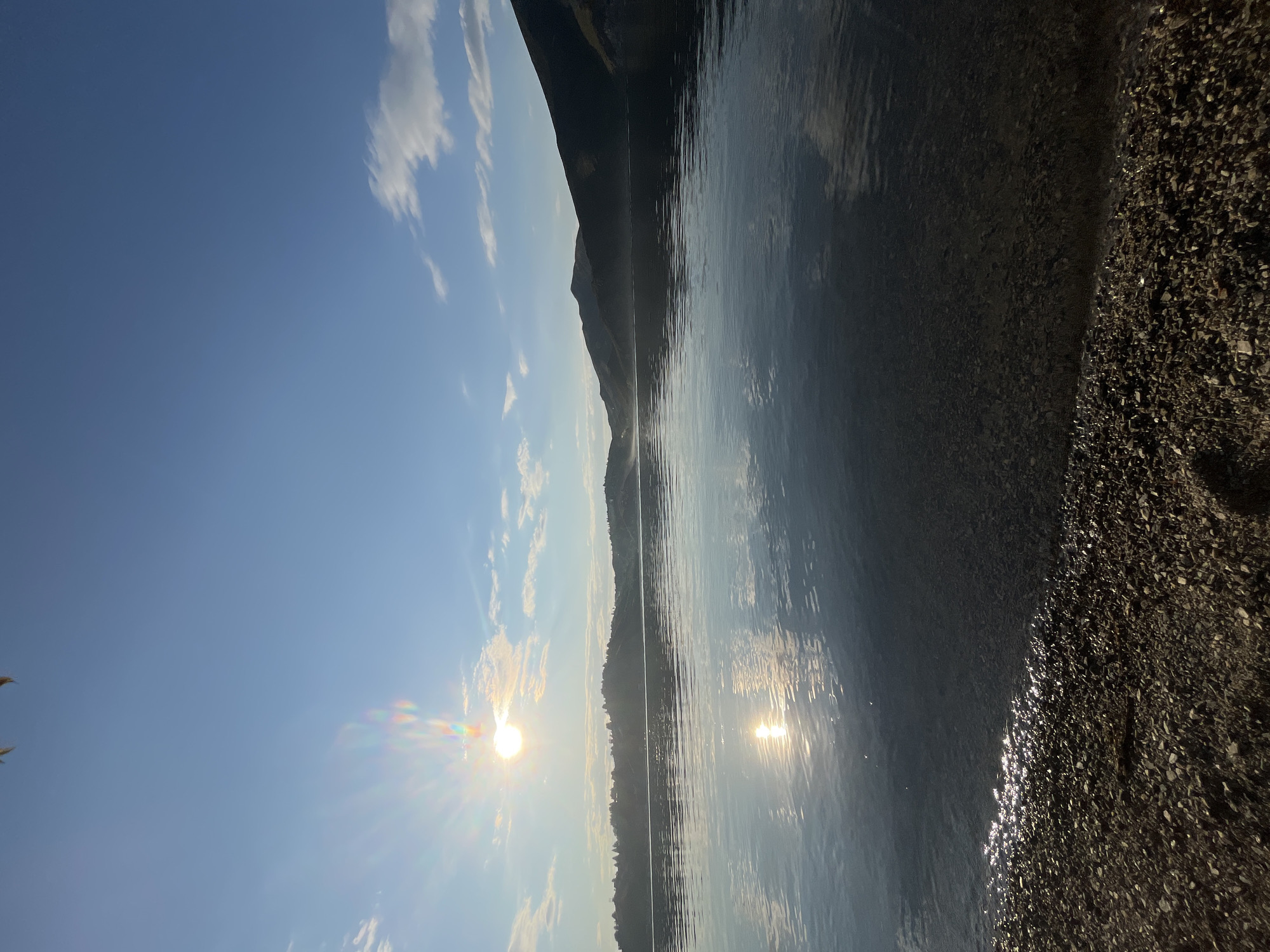 This is an eye-level, full shot photograph of a lake with mountains in the background. The sun is setting or rising over the mountains, casting a bright glare on the water. The sky is a clear blue with a few wispy clouds. The water is calm and reflects the sky and the mountains. The shore in the foreground is covered with small rocks. The scene is peaceful and serene. The location is the Irtysh River near the city of Oskemen, Kazakhstan.