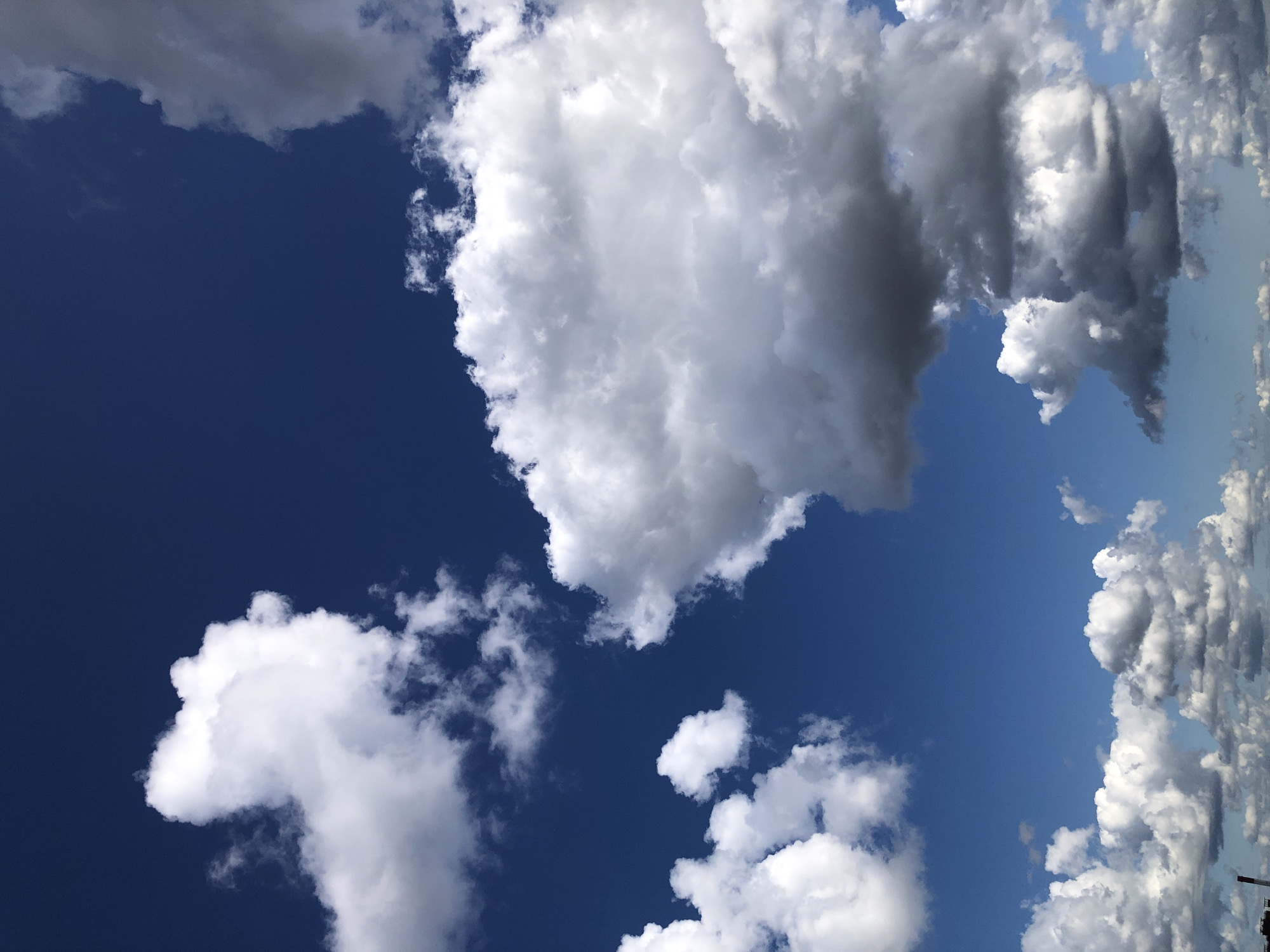 An outdoor, eye-level shot captures a deep blue sky filled with a variety of white and gray clouds. The clouds are predominantly cumulus, with some appearing puffy and bright white, while others are larger and darker gray, suggesting greater density or depth. The lighting indicates it is daytime, with the sun illuminating the upper portions of the clouds. No people or specific man-made structures are prominently visible, except for the very top of what appears to be a smokestack or industrial chimney on the lower left edge of the frame, hinting at an urban or industrial setting. The overall scene is natural, focusing on the atmospheric conditions.
