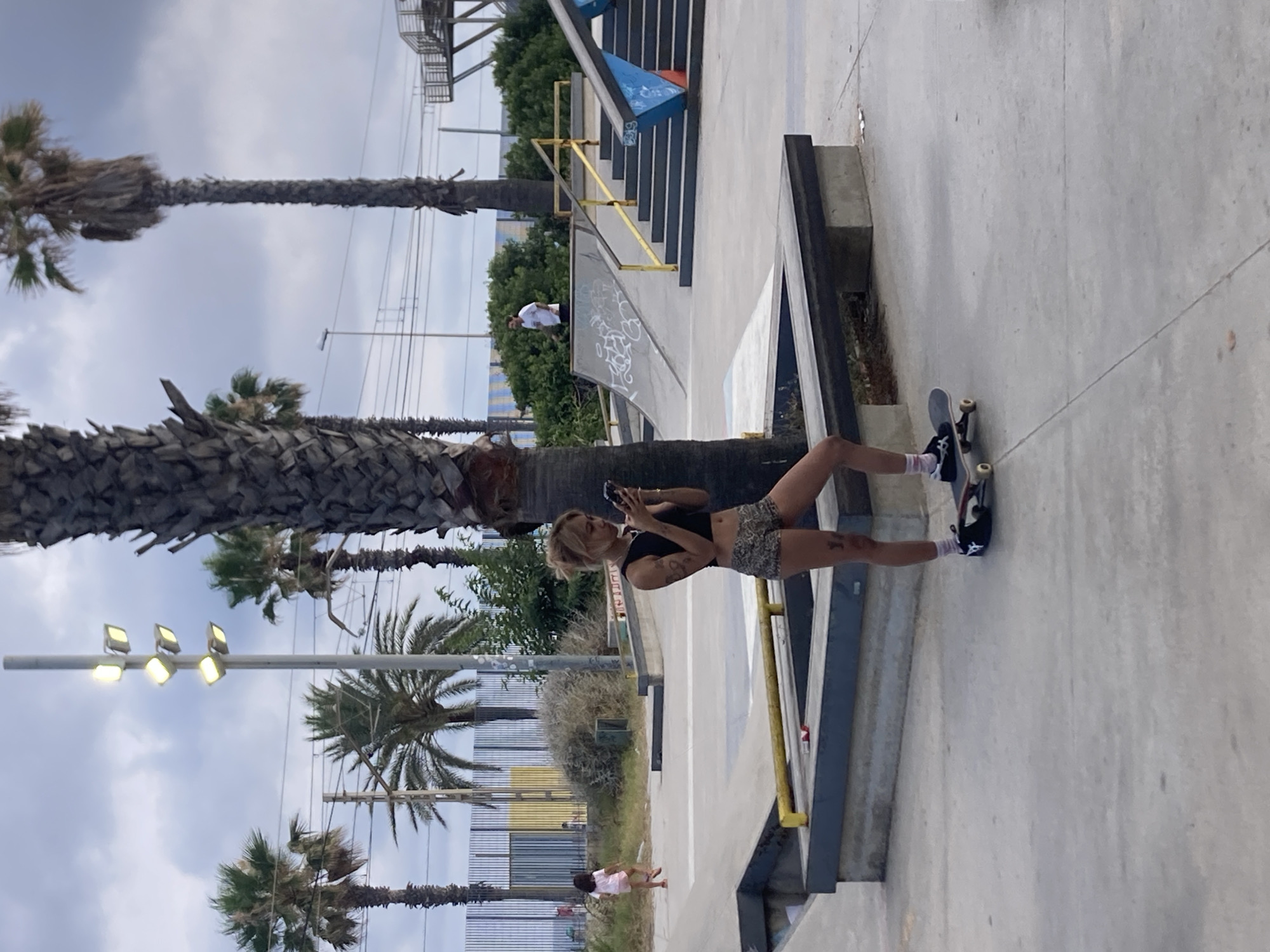 The image depicts a young woman standing on a skateboard in a skatepark, holding a smartphone. She is wearing a black crop top and patterned shorts. The skatepark features ramps, ledges, and palm trees, indicating a warm climate. In the background, a small child can be seen running, and further in the distance, a man is standing near a ramp. The sky is overcast, suggesting a cloudy day. The location is identified as the skatepark in Badalona, Spain.