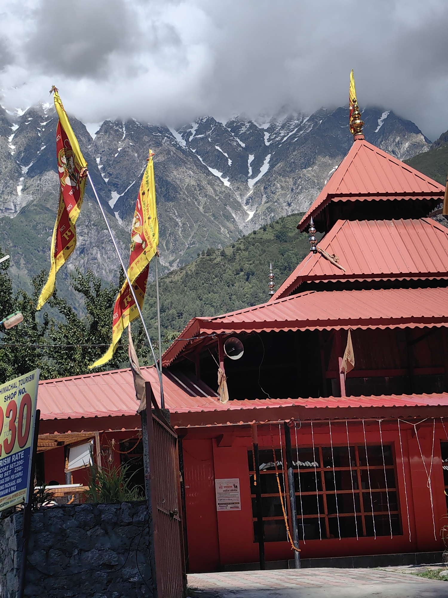 In the city of Kulang, India, a brightly painted red temple stands before a backdrop of majestic, snow-capped mountains. The temple's tiered, red-tiled roof is adorned with decorative finials. In front of the temple, two vibrant yellow and red prayer flags flutter in the breeze, bearing religious inscriptions. A sign indicating "HIMACHAL TOURISM SHOP NO. 30" is visible to the left, providing a glimpse of local commerce. The scene is further framed by lush green trees and a rustic stone wall. The sky above is partially obscured by dramatic, dark clouds, adding a sense of grandeur to the mountainous landscape.