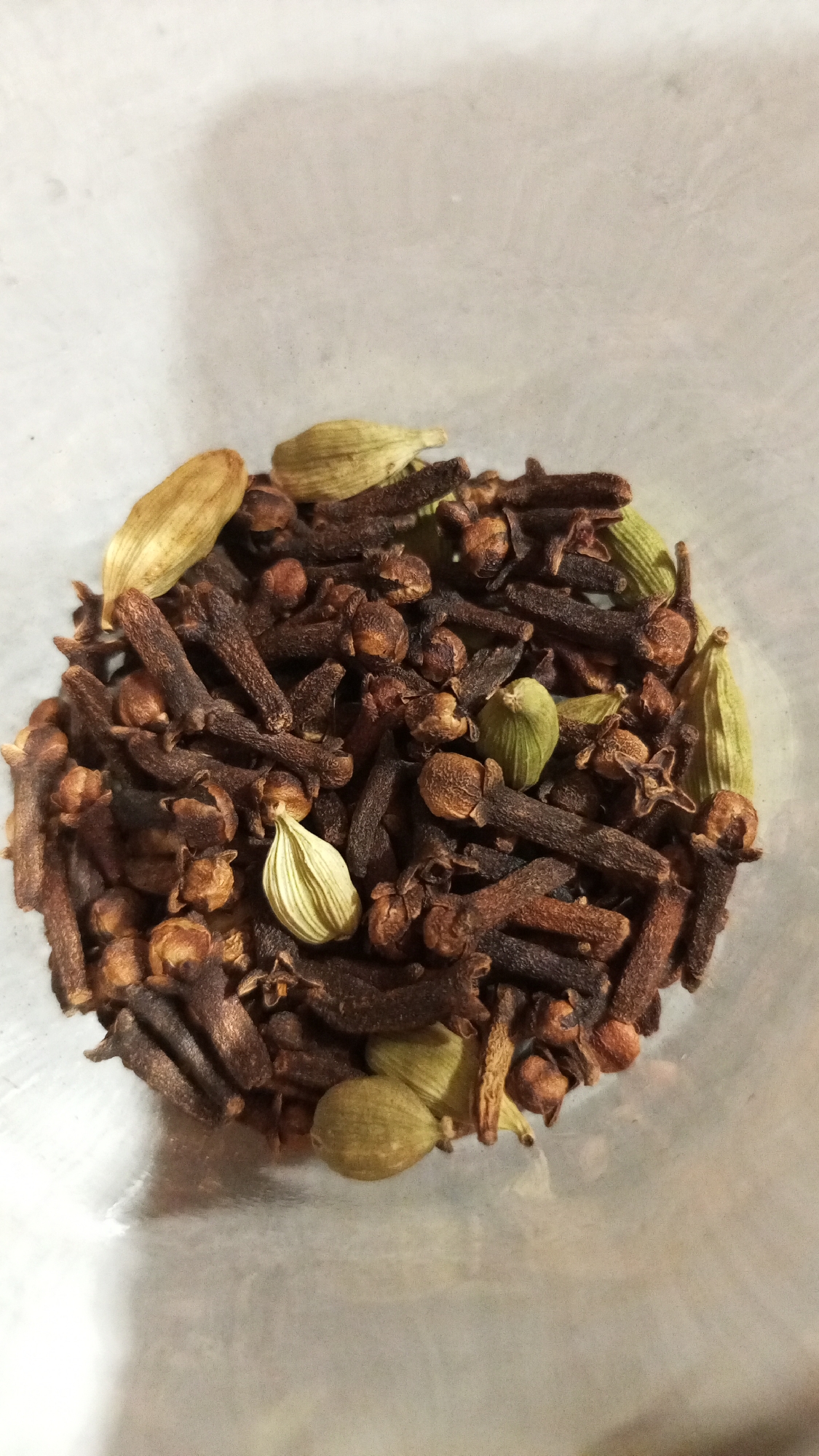 The image shows a close-up, top-down view of a collection of cloves and cardamom pods. The cloves are predominantly dark brown, with a distinctive stem and a rounded head. The cardamom pods are pale green to light brown, with an elongated, oval shape. They are interspersed amongst the cloves. The objects are contained within a clear, possibly glass or plastic container. The background is a slightly blurred, light gray, likely indicating the surface upon which the container rests. The location Юдино, Russia is extraneous to the contents of this image.