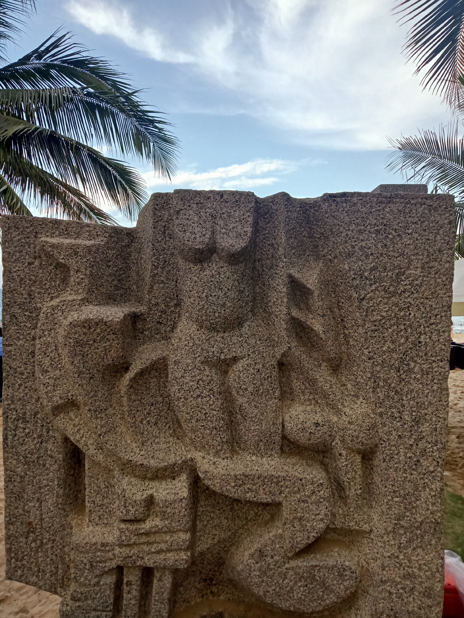 The image shows a large, rough-hewn stone sculpture in front of a beach. The sculpture appears to be carved with abstract figures, possibly depicting people or deities. Palm trees are visible in the background, along with a sandy beach and the ocean. The sky is partly cloudy. The location is in Nha Trang Ward, Vietnam.