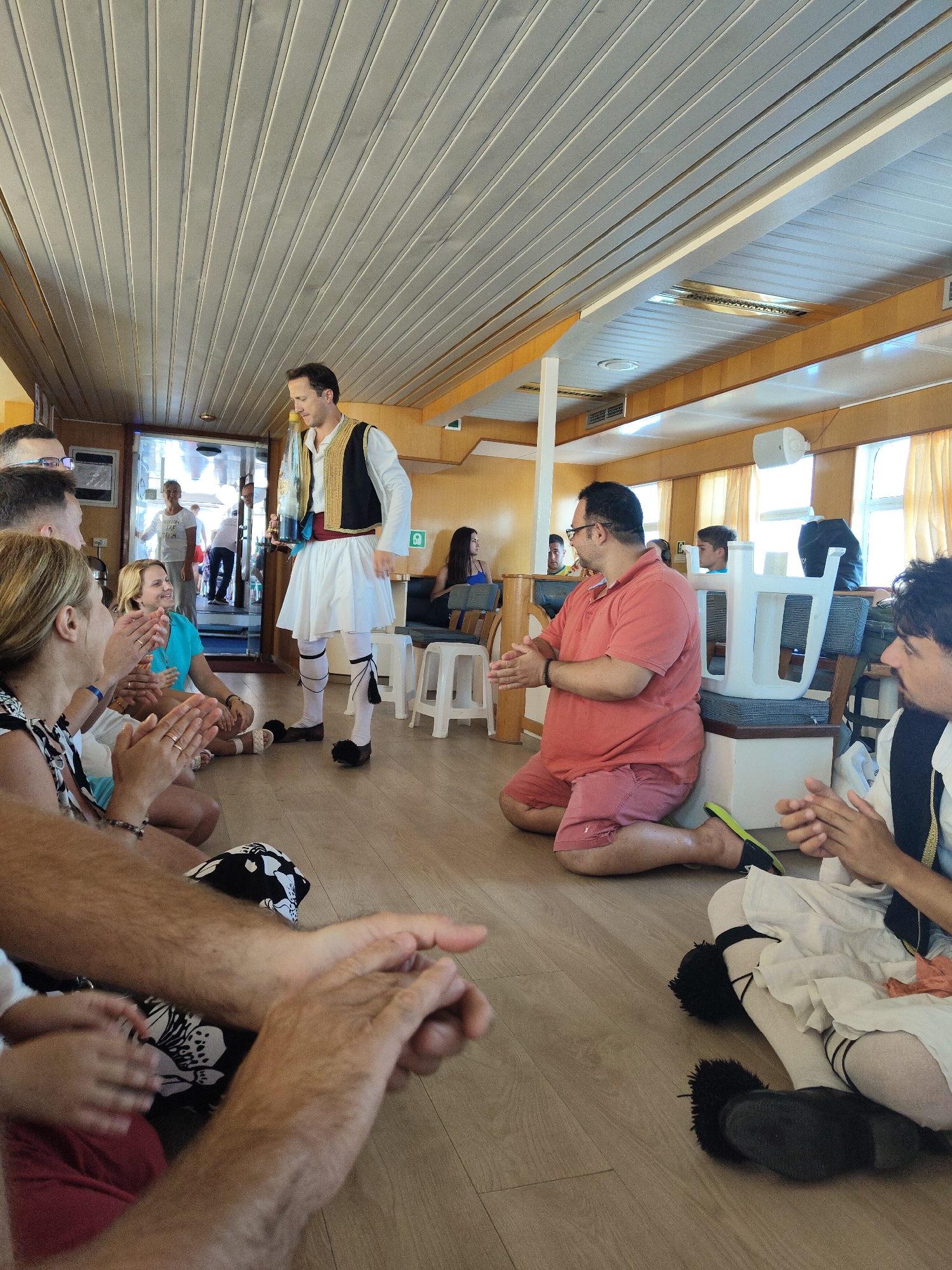 The image depicts an indoor scene, likely on a boat or a venue with a wooden paneled ceiling and wooden floor. The location is described as Unknown, Greece. A man dressed in traditional Greek attire, including a white fustanella kilt, a black vest adorned with gold trim, and pom-pom adorned shoes, stands in the center of the room. He is holding two bottles, possibly for a demonstration or performance.
A group of people are seated on the floor around him, many of them clapping. The audience appears to be a mix of ages and genders, with some individuals wearing casual clothing. One woman in the foreground is wearing a black and white patterned top and red shorts, and she is clapping with her hands held together. Next to her, another woman in a turquoise top is also clapping. To the right, a man in a coral-colored polo shirt and red shorts is kneeling on the floor, also clapping. Further to the right, another man is seated, clapping.
In the background, several people are standing near an open doorway, one woman in a white t-shirt is visible. The room is lit by natural light streaming through windows with yellow curtains and by overhead lighting. White stools are visible in the background, suggesting a seating area. The overall atmosphere appears to be festive and engaged, with the audience actively participating in the event.