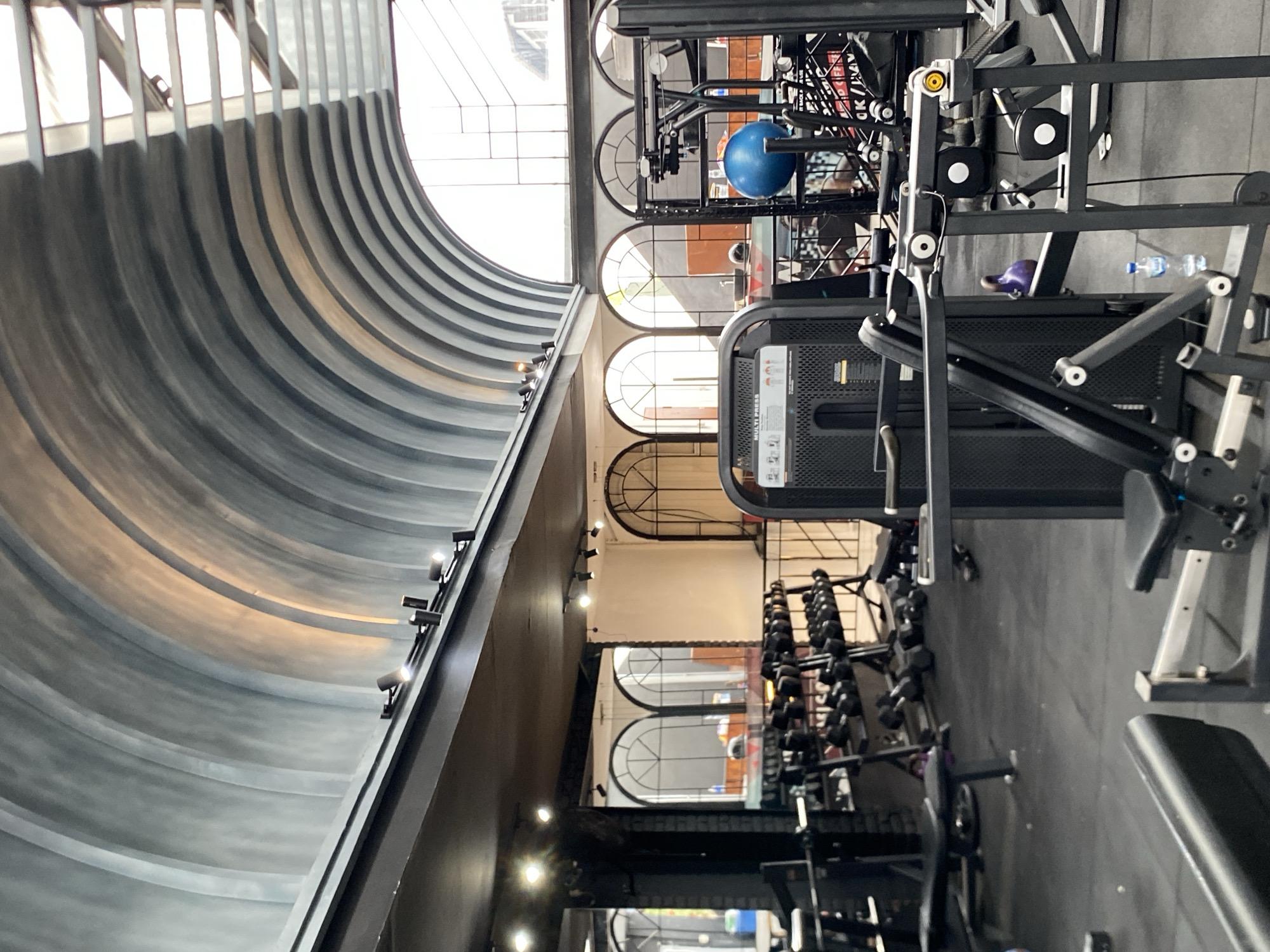 This is a wide-angle, indoor photograph of a gym in North Kuta, Indonesia. The gym features a distinct arched ceiling with a ribbed texture, painted in shades of grey. The ceiling has multiple track lights directed downwards. Below the ceiling, a dark-colored overhang runs horizontally across the image. The gym floor is black and rubberized.
Dominating the foreground are various pieces of weight training equipment, including a multi-press machine on the left, and other cable machines. A kettlebell, colored purple, is visible on one of the machines. There is a bottle of water on the floor near the machines.
In the mid-ground, a rack of dumbbells is neatly arranged. Behind the dumbbells, arched windows with decorative metalwork are visible, reflecting light from the outside. Mirrors are mounted on the wall behind the dumbbell rack, reflecting the arched windows and parts of the gym.
In the background, through the windows and in the reflected areas, an outdoor space with some greenery and what appear to be buildings can be seen. A person is partially visible in the background, seated, with a blue exercise ball on a shelf behind them. The overall impression is a modern, well-equipped gym with an emphasis on architectural design in its ceiling structure.