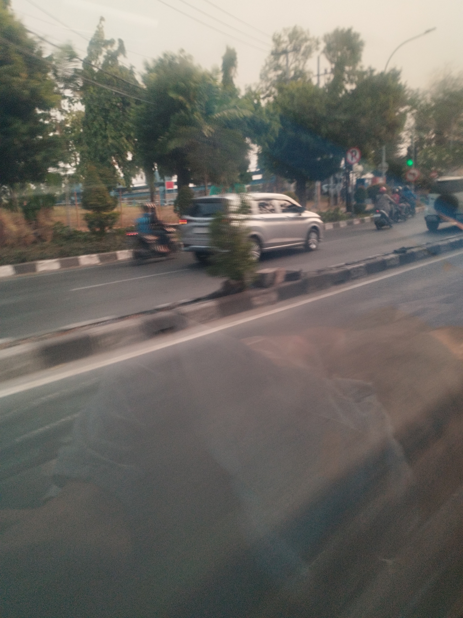 The image captures a blurred street scene in Surabaya, Indonesia, characterized by a mix of vehicular and pedestrian activity under an overcast sky. A silver SUV is visible on the right side of the road, appearing to be in motion. To its left, several motorcycles are also present, with riders wearing helmets and casual attire. The road itself is paved, with a distinct white line marking the lane and a raised curb on the side. Beyond the road, a row of trees and some buildings can be seen, suggesting an urban environment. The overall impression is that of a typical day on a busy street in Surabaya, with the motion blur indicating movement captured by the camera.