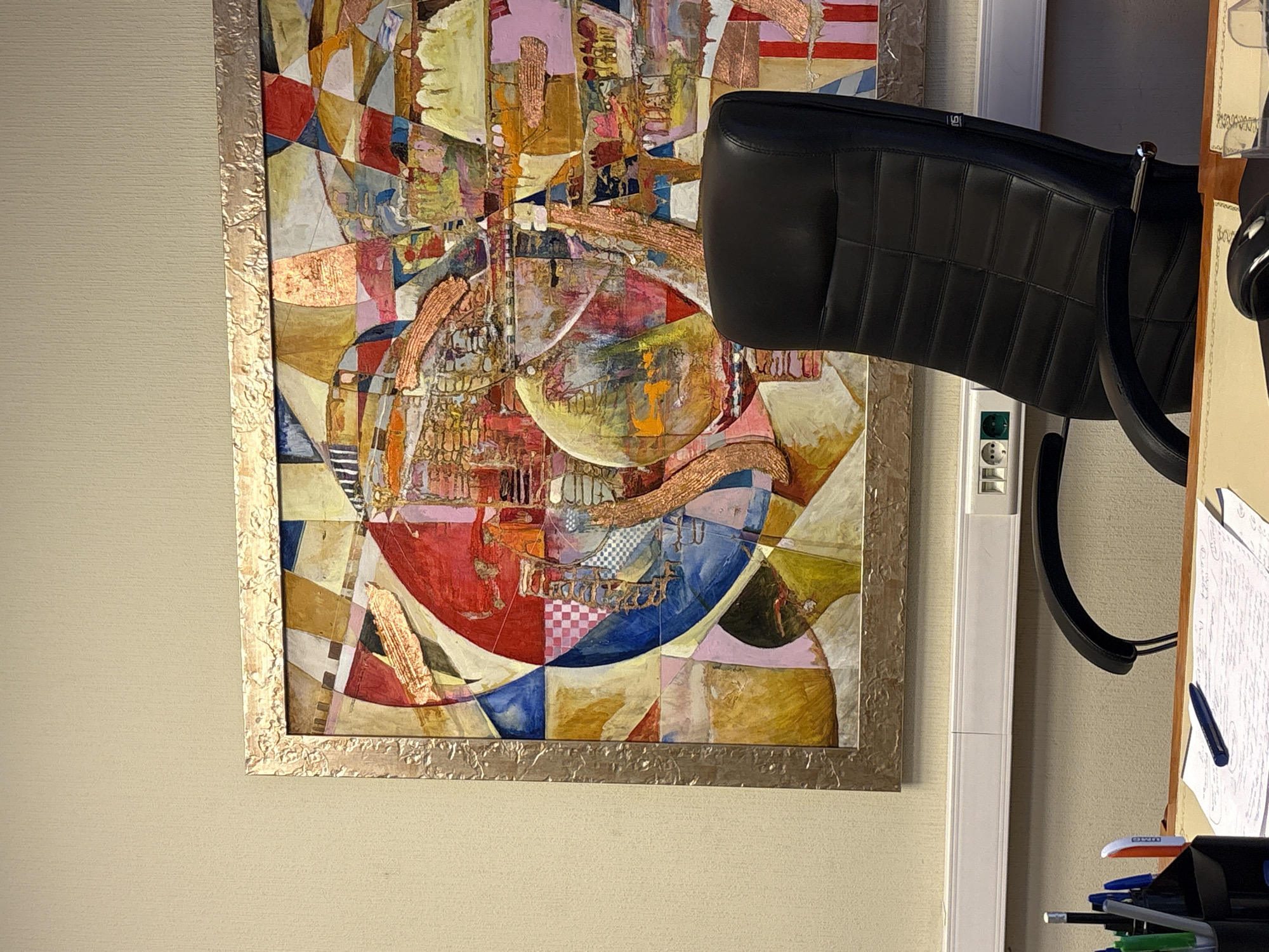 The image shows an interior scene. On the left is a wall with a framed abstract painting, featuring a patchwork of geometric shapes in various colors, including red, blue, yellow, pink, and white. The frame is gold-colored and appears textured. To the right of the painting is a white vertical structure, possibly a cable runner or conduit. A black leather chair with horizontal stitching is positioned in the right foreground, partially obscuring the white structure and the painting. Further to the right, there's a table or desk with papers, pens, and other office supplies. The environment suggests an office or study setting. There is no information in the image about Moscow or Russia.