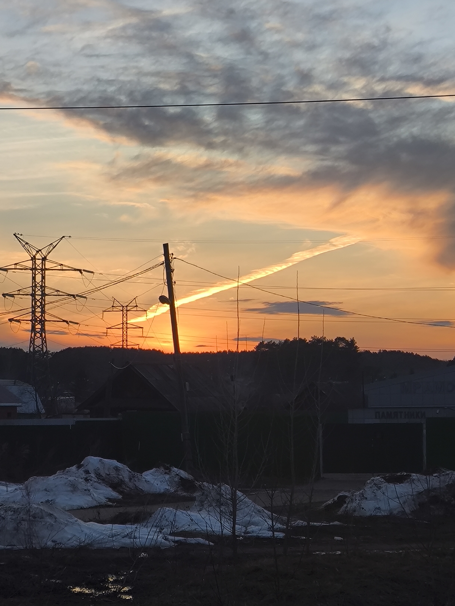 The image depicts a sunset scene in what appears to be a residential area near the city of Chaikovsky, Russia. The sky is a mix of orange, yellow, and gray clouds, with a contrail visible in the upper part of the frame. In the mid-ground, there are electrical power lines, utility poles, and the silhouettes of houses and trees. The lower portion of the image shows snow-covered ground and bare vegetation, suggesting late winter or early spring. The buildings in the background have signage with Russian text. Overall, the image conveys a sense of a peaceful, rural or suburban setting at dusk.