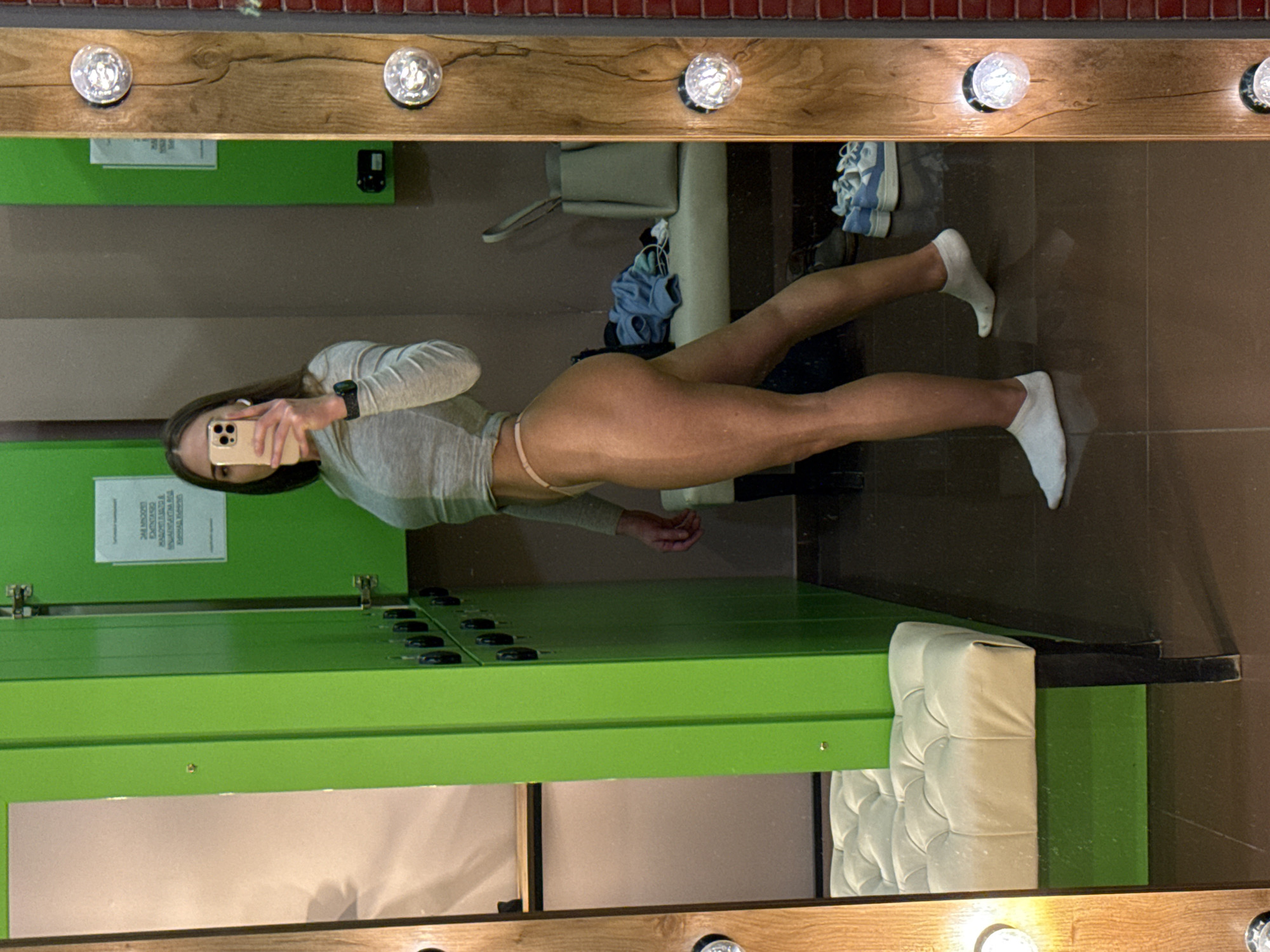 An image captures a woman taking a mirror selfie in what appears to be a changing room or locker room in Moscow, Russia. The setting features bright green lockers on the left, a light-colored tufted bench on the right, and a dark tiled floor. The mirror, framed by light brown wood with several round, clear light bulbs, reflects the scene.
The subject, a young woman with long dark hair, is positioned to showcase her physique. She wears a light gray long-sleeved crop top, light pink thong-style underwear, and white athletic socks. She holds a light-colored smartphone (an iPhone) to take the photo, with white wireless earbuds in her ears and a dark smartwatch on her left wrist. Behind her, light blue and white sports shoes and some scattered clothing items are visible on the floor.
A white paper sign is attached to one of the green lockers. Although partially obscured and angled, discernible text includes "24 ЧАСОВЫЙ ДОСТУП" (24-HOUR ACCESS) and "ПРАВИЛА ПОСЕЩЕНИЯ" (RULES OF VISIT), indicating the facility's operational hours and guidelines. The overall impression is that of a fitness or gym changing area, likely indoors, with artificial lighting provided by the mirror's bulbs.