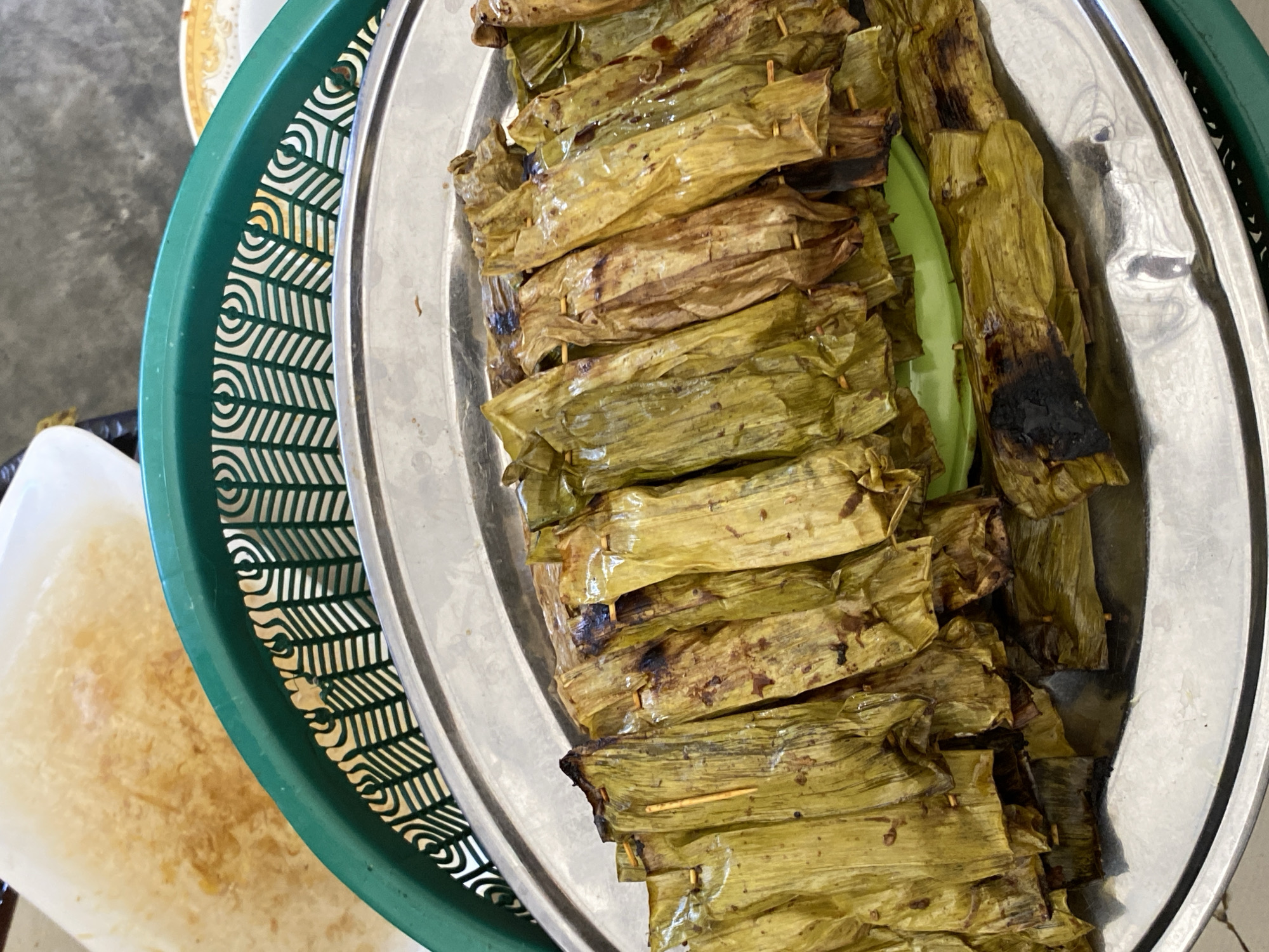 A close-up, overhead shot shows a platter of grilled food wrapped in banana leaves. The food items, each approximately 4-5 inches long, are arranged in a neat row on a large, round, silver platter. They appear to be secured with small wooden skewers. The banana leaves are slightly browned and charred in places, indicating they have been grilled or cooked over heat. Some of the leaves are a vibrant green, while others are a muted yellow-brown. In the background, a green plastic basket with a decorative pattern is visible, and behind that, a white and yellow patterned surface, possibly food or a serving tray. The setting appears to be outdoors or in a casual dining environment, consistent with street food stalls in a city like Banda Aceh, Indonesia, where such dishes are commonly prepared.