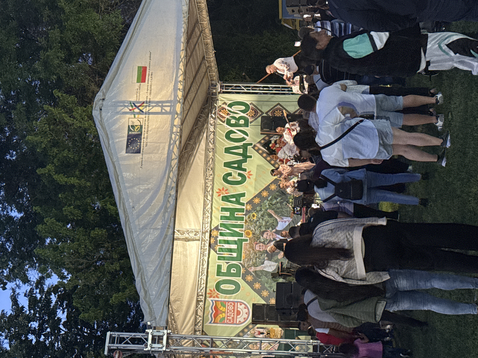 The image depicts an outdoor stage set up for a performance in Sadovo, Bulgaria. A crowd of people is watching the performers on stage. The stage is covered with a white tent structure, and a large banner with the words "ОБЩИНА САДОВО" (Municipality of Sadovo) is visible on the stage backdrop. The banner also features images of sunflowers and people. A Bulgarian flag and the EU Leader program logo are displayed. Trussing is visible on the stage. The environment appears to be a park or open space, with trees in the background.