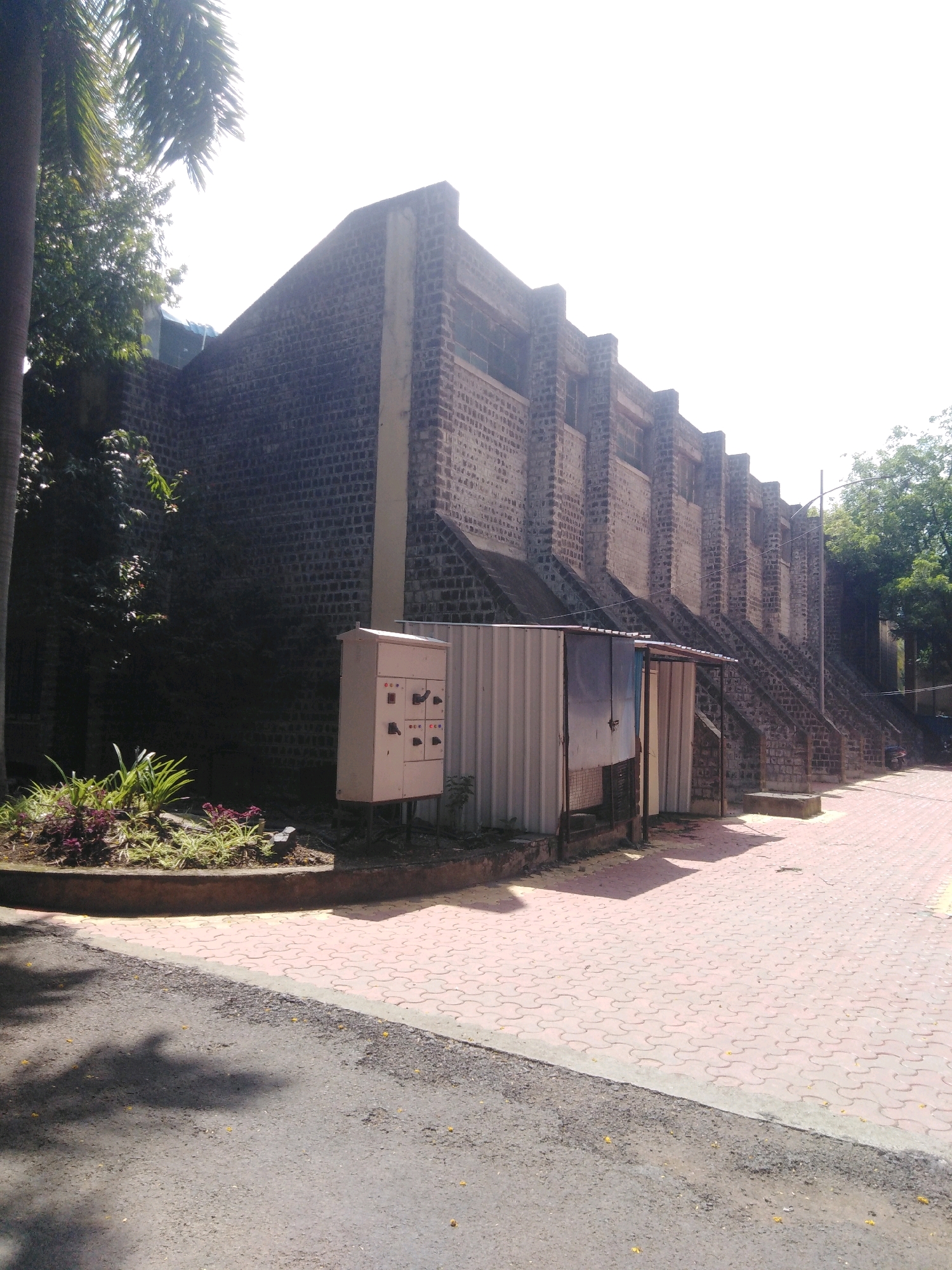 The image shows the side of a brick building with a sloping roof and a series of buttresses. In front of the building, there is a paved walkway with interlocking red bricks, and a road made of asphalt. There are some trees and plants visible, as well as an electrical panel box and a small shed with a metal roof. The scene is illuminated by bright sunlight, casting shadows on the ground. This image is likely from the campus of Swami Ramanand Teerth Marathwada University in Nanded, India, based on similar architectural styles and landscaping found in the area.